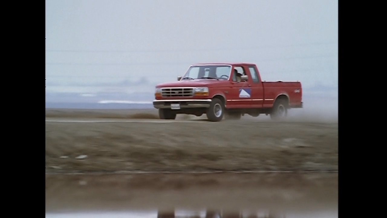 A red pickup truck drives along a dirt path bordering a reflective body of water, followed by a man operating a valve in the adjacent area.
