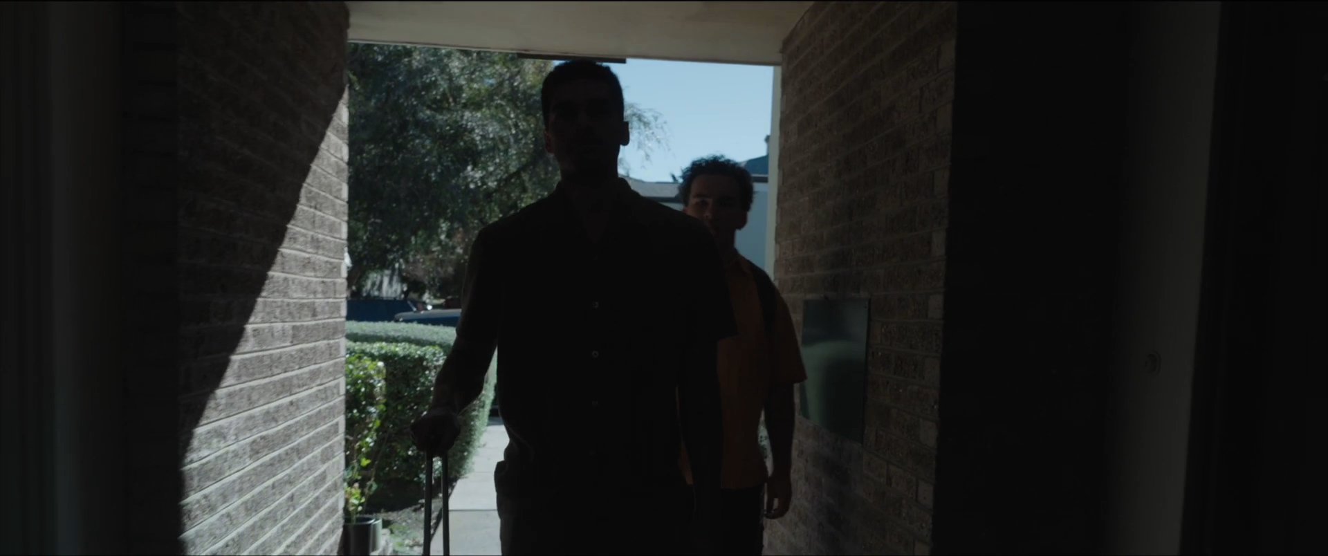 Two men arrive at a residence carrying luggage and enter the living room, where a friend watches television.
