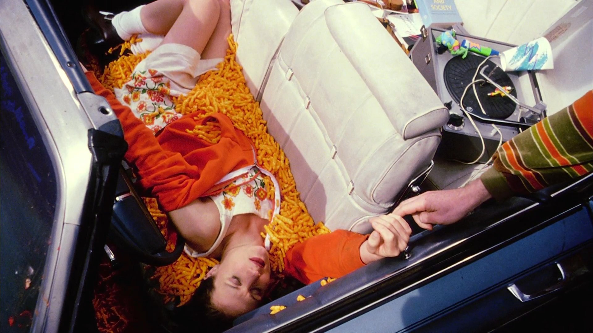 A hiker wearing a large backpack approaches a woman reclining in a convertible filled with snack food in a grassy field. He offers a hand to help her exit the vehicle while they share a brief, intimate interaction.
