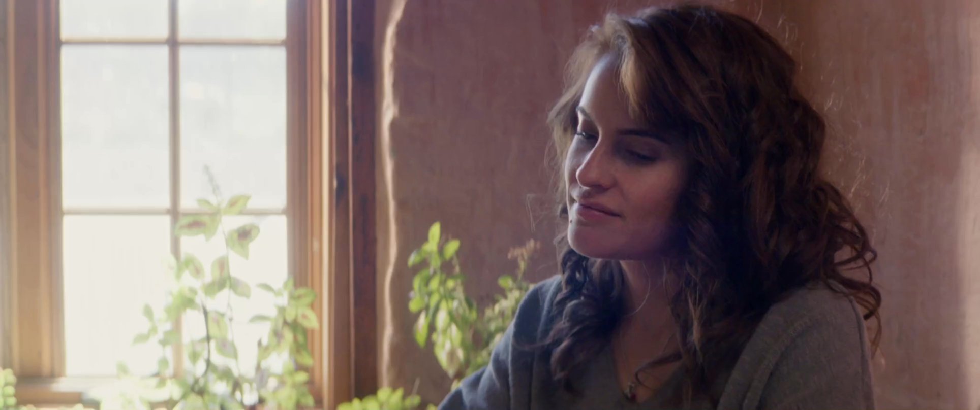A man and a woman engage in a quiet, intimate conversation while seated at a wooden table near a sunlit window.