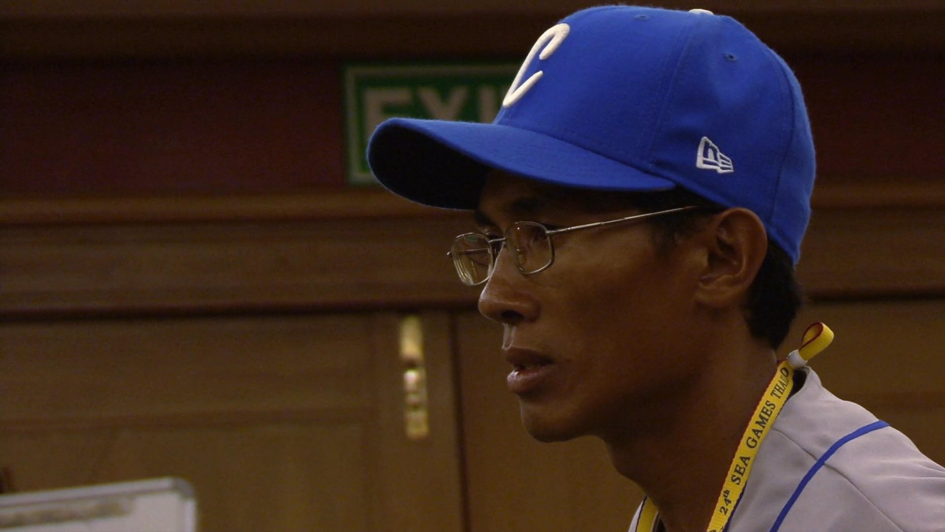 A coach wearing a baseball uniform addresses a group of reporters during an organized press conference. He answers questions and gestures to explain team strategy and tournament expectations.