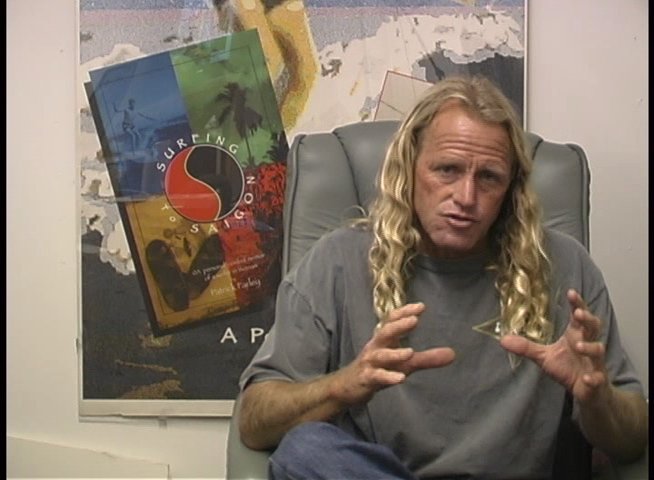 A long-haired professional surfer sits in an office setting, actively discussing experiences and insights through expressive hand gestures.