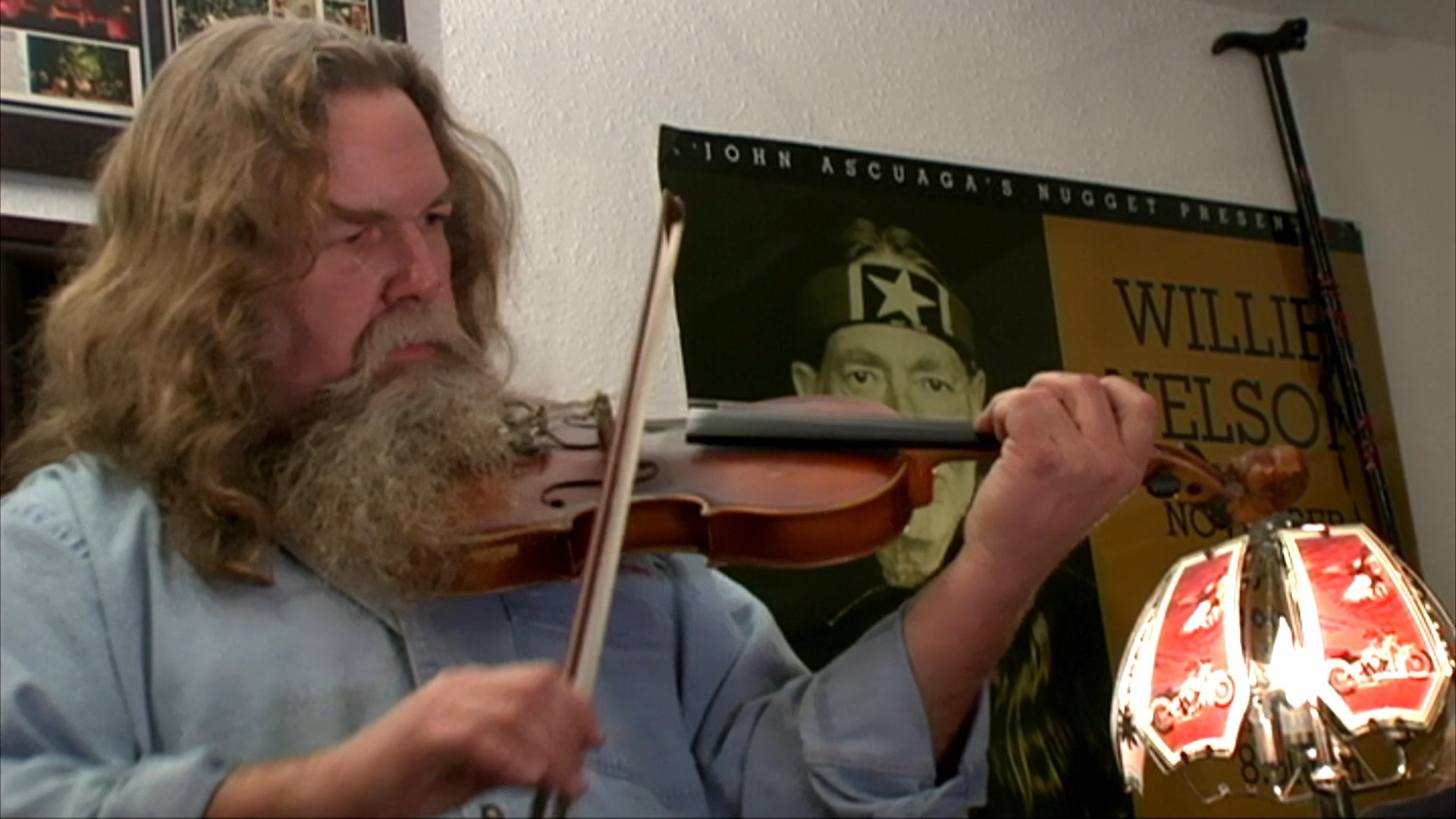 A long-haired, bearded man plays the violin in an indoor setting before the footage shifts to nostalgic home movie clips of a child playing outdoors and a family dinner.