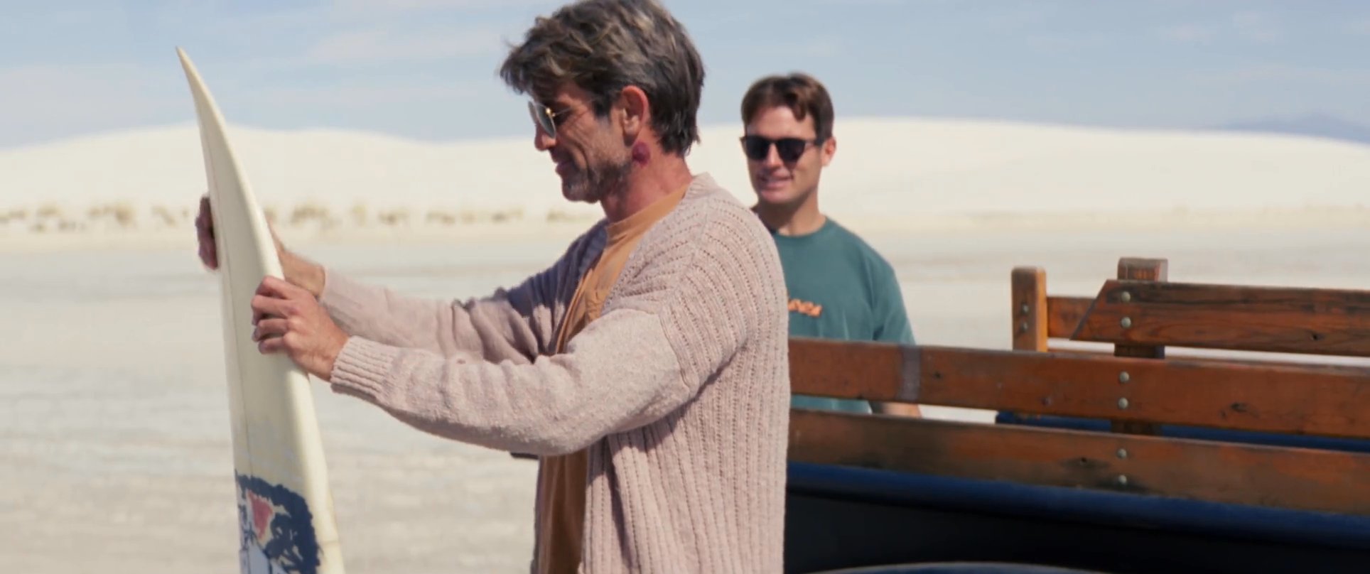 Two individuals retrieve surfboards from the back of a vintage pickup truck before trekking across expansive white sand dunes.