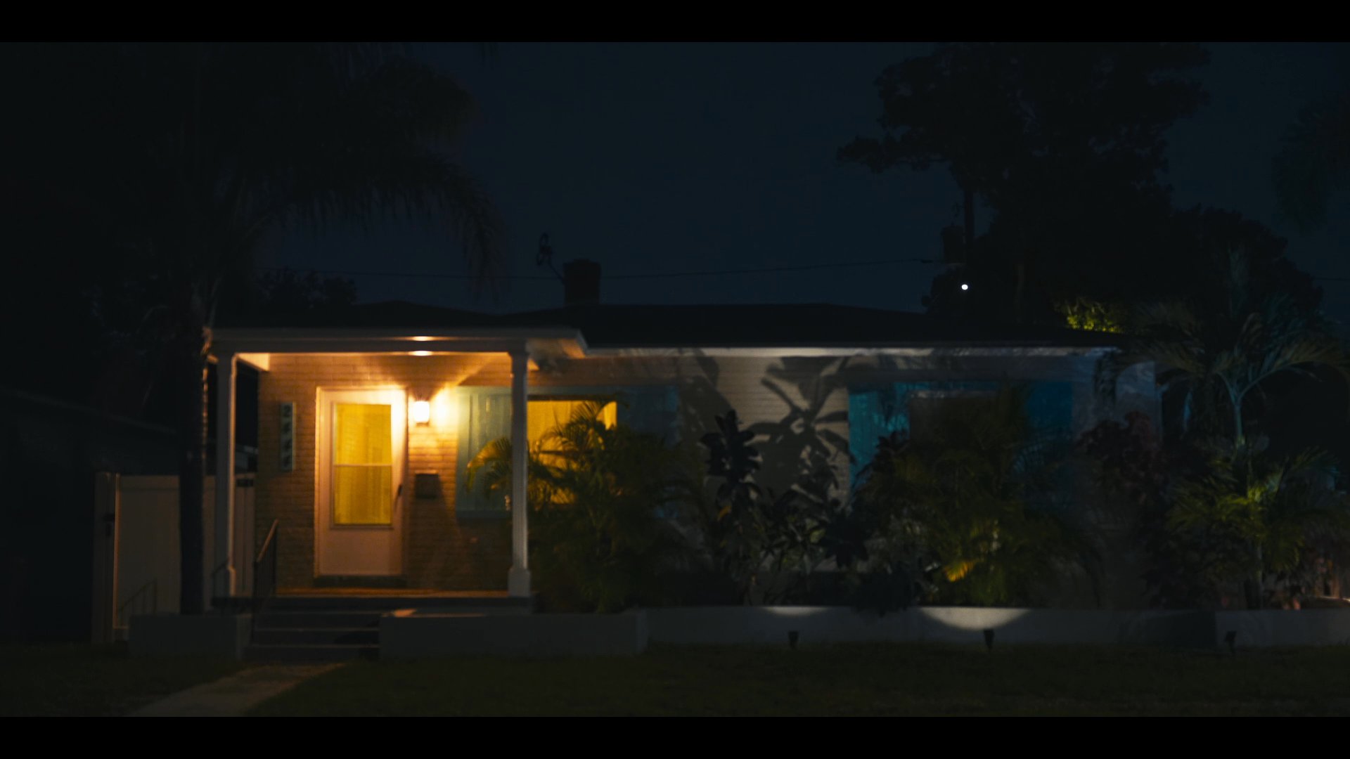 A man walks through a sticker-covered bar area, followed by a cut to an exterior shot of a house at night, ending with a man lying in bed and speaking on his phone.