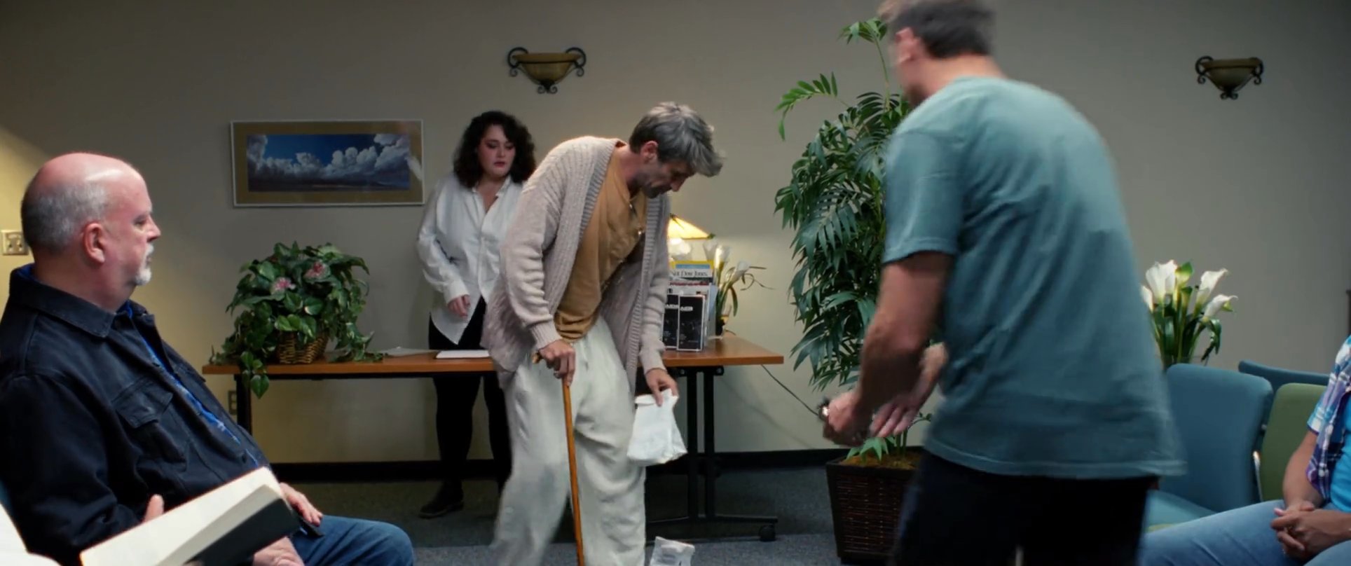 An injured man with a cane attempts to navigate a tense environment while interacting with others in a quiet, professional waiting room. The interaction highlights a moment of social discomfort and physical struggle as he moves through the shared space.