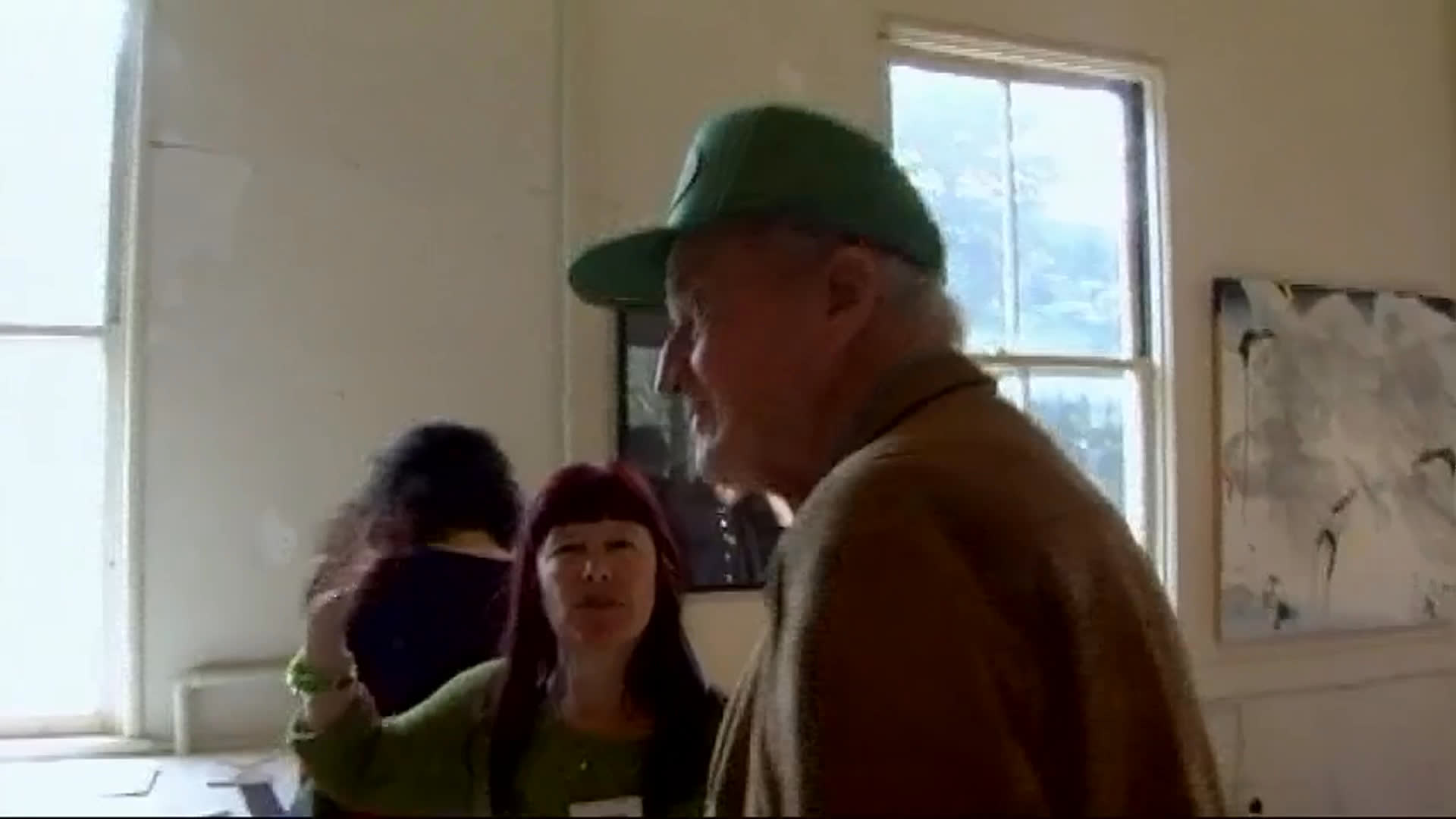 An elderly man in a green cap moves through a bright gallery, studying artworks and speaking with visitors. He expresses admiration for a piece and describes art as his primary source of financial engagement.