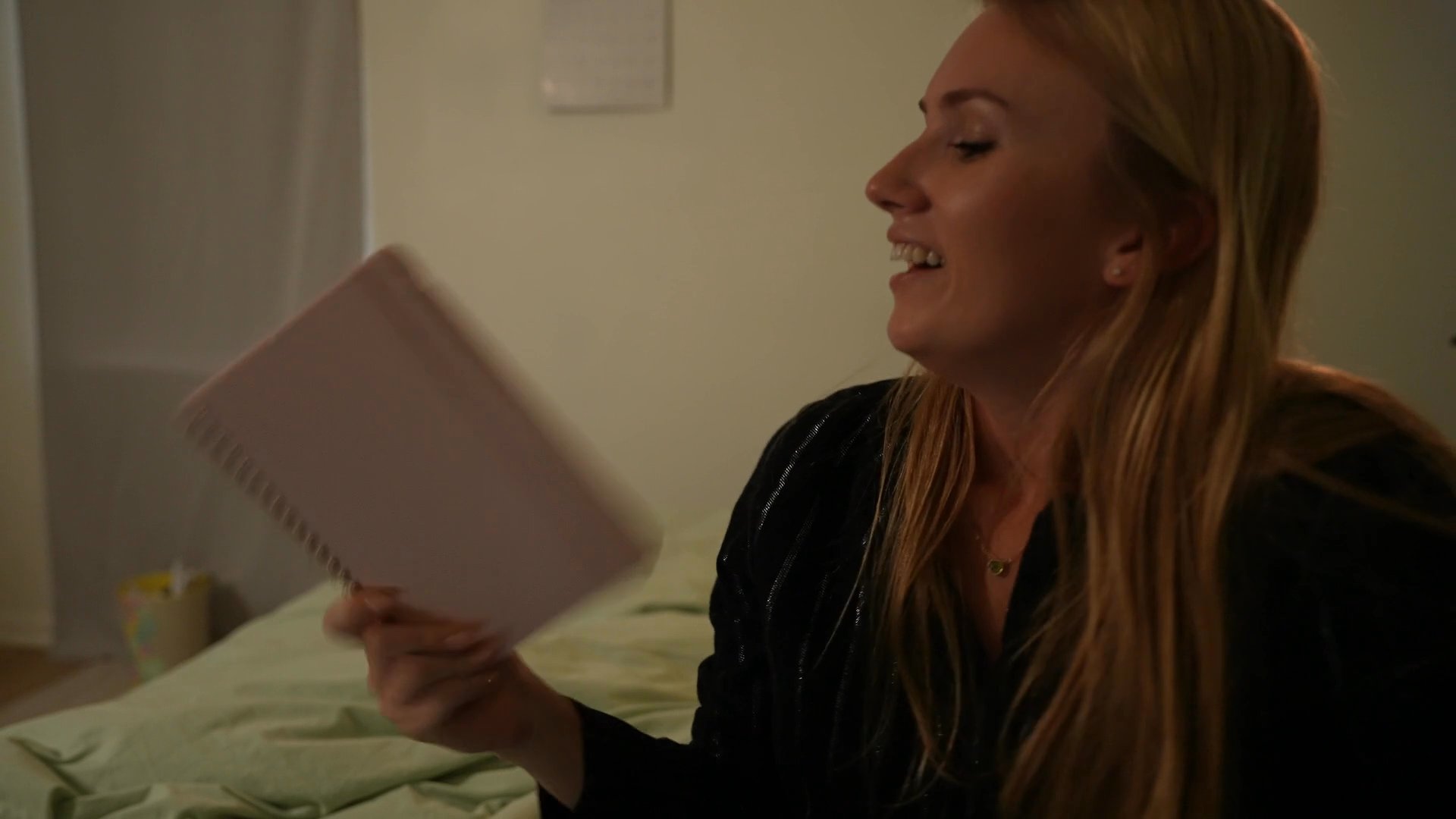 A young woman sits on her bed, alternating between drinking from a mug and laughing while looking at a notebook. The scene captures a private, lighthearted moment of rest in an evening bedroom setting.
