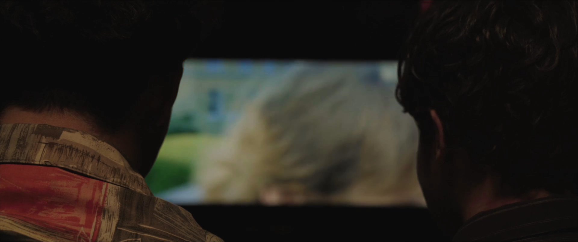 Two individuals sit in a darkened theater, watching a film projection while periodically exchanging expressions and reactions.