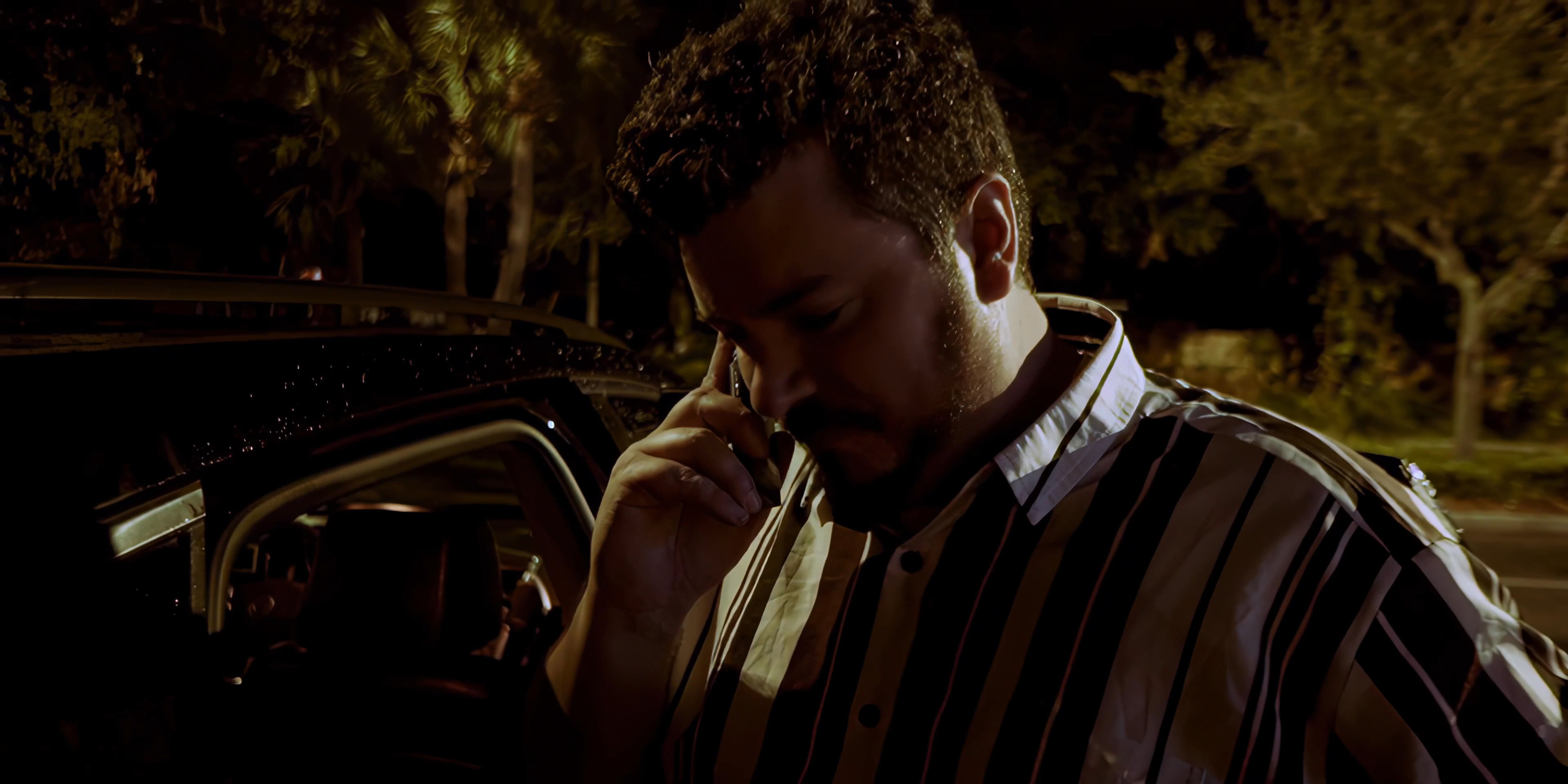 A man in a striped shirt makes a serious phone call while standing outside next to a car at night.
