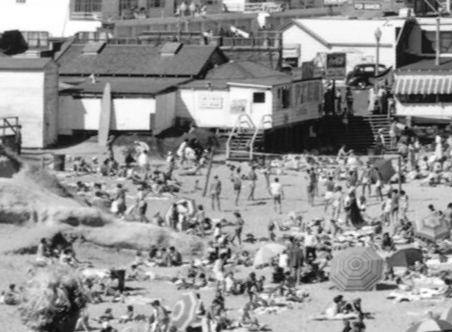 Crowds of people enjoy a sunny day at a populated beach set against a backdrop of coastal structures. The view shifts from a wide perspective of the beachgoers to a closer look at individuals observing the scene from a nearby vantage point.