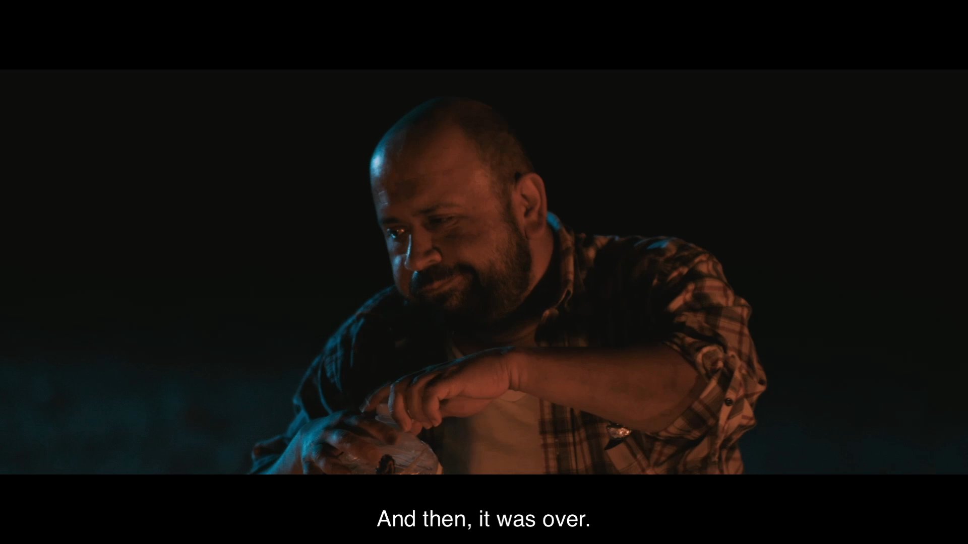 Two men exchange personal stories beside a campfire at night, with one recounting a past incident involving wood planks and an encounter with the police.