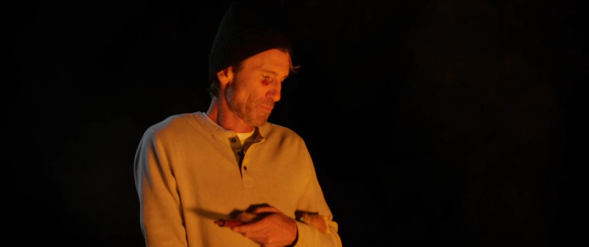A man addresses a small group gathered around an outdoor fire pit at night. The warm glow of the flames illuminates the scene as the group enjoys a quiet, intimate evening together.