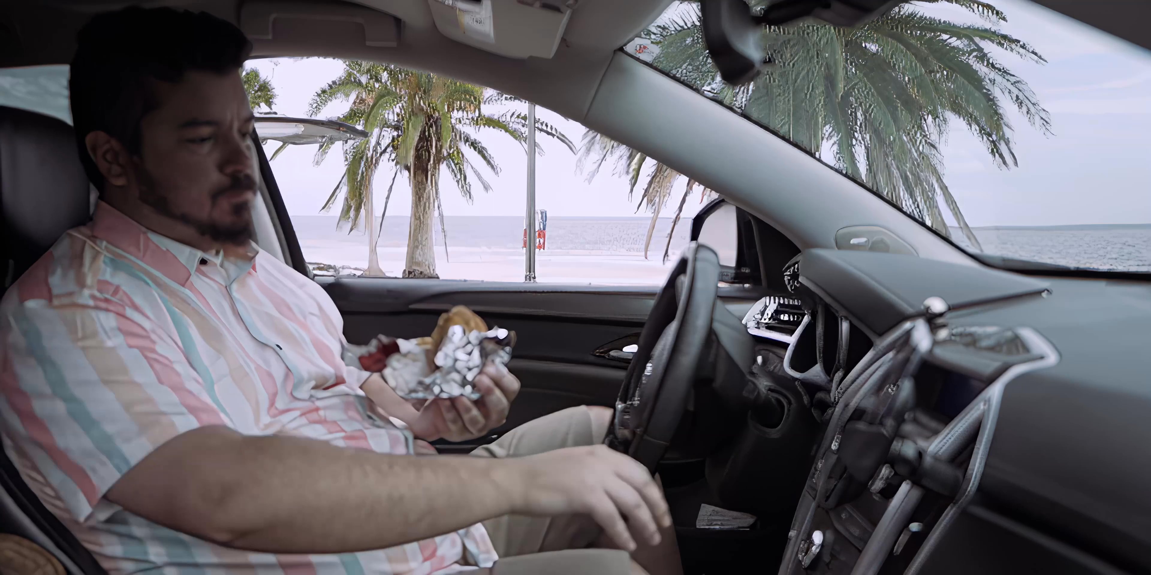 A man sits in a parked car by the beach, unwrapping and eating a hamburger. He pauses to interact with the vehicle's infotainment system before continuing his meal.