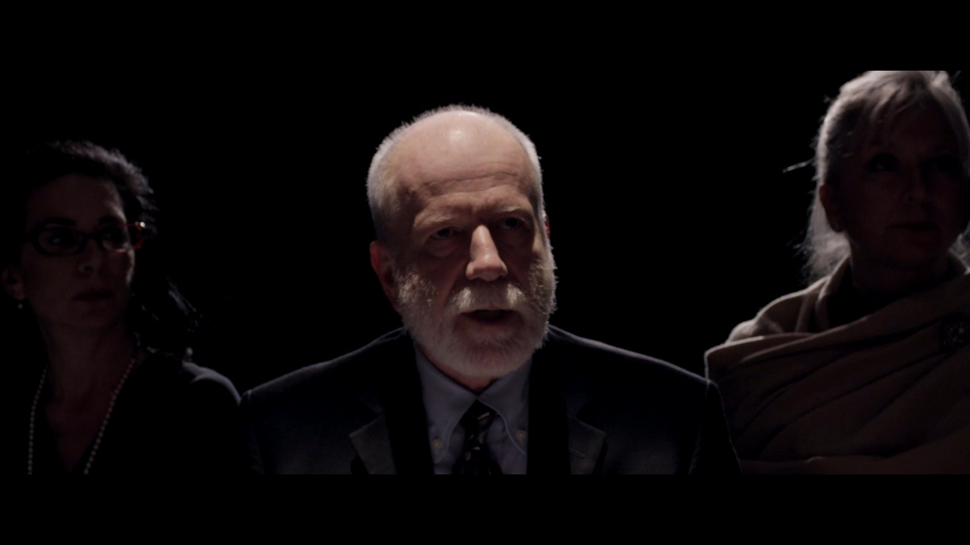 A group of individuals participate in a formal casting session, showcasing a range of subtle expressions and reactions against a dark background. The scene captures the tension and focus typical of a professional theatrical audition.