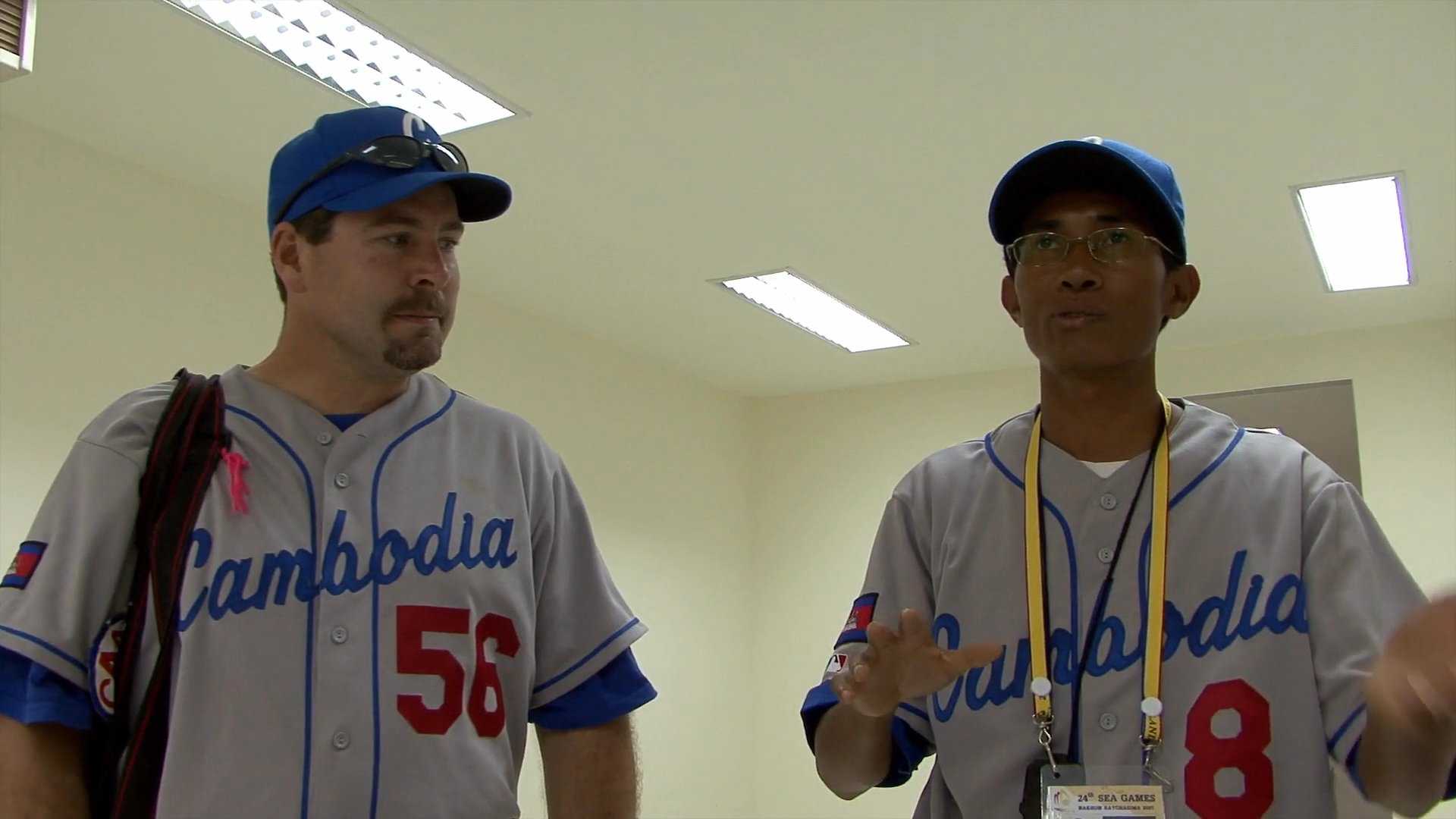 Two individuals wearing Cambodia baseball uniforms stand together in an indoor facility and engage in a focused, professional conversation.