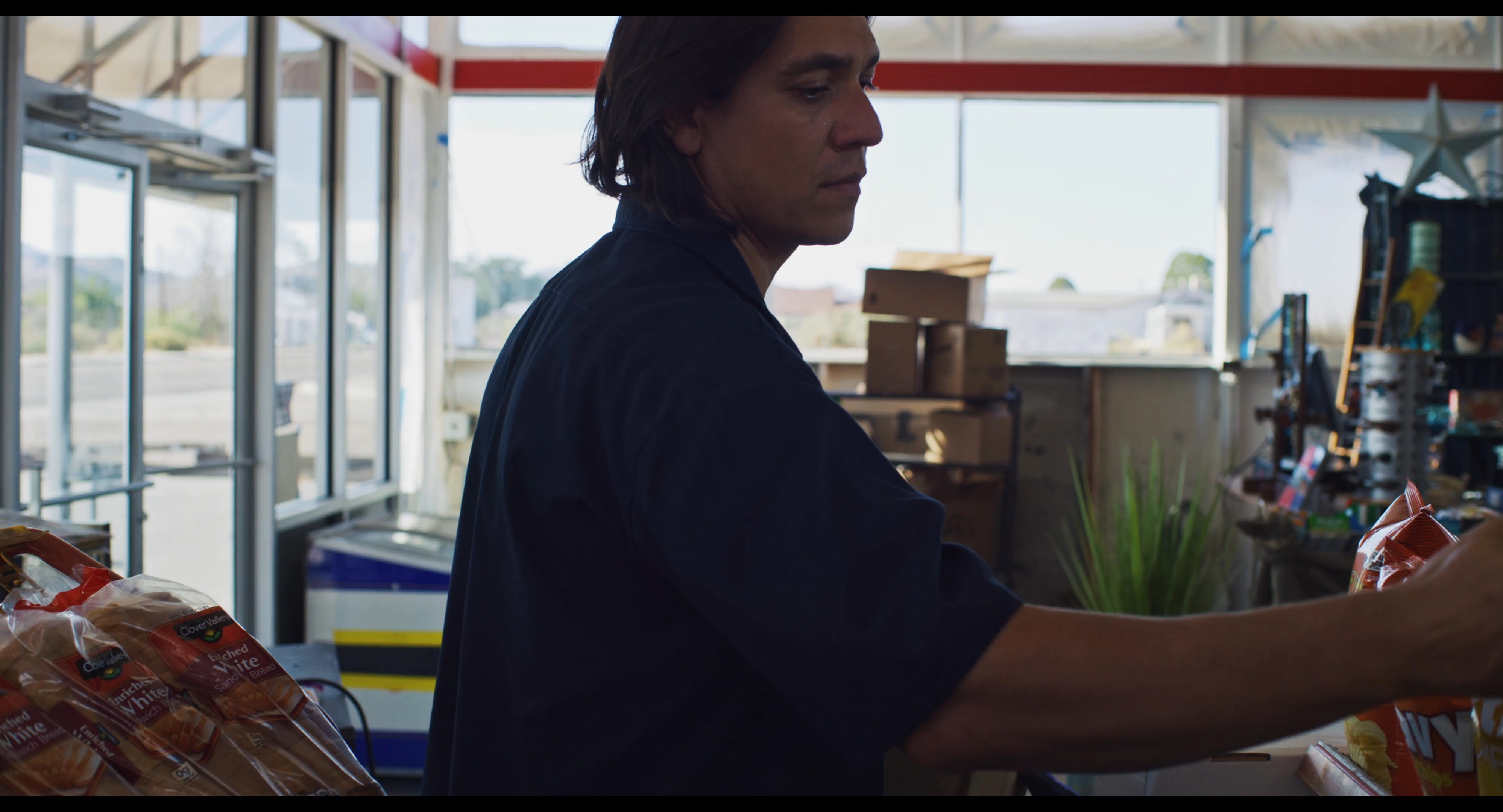 A store clerk organizes merchandise on shelves within a rural roadside convenience store while customers navigate the interior space.