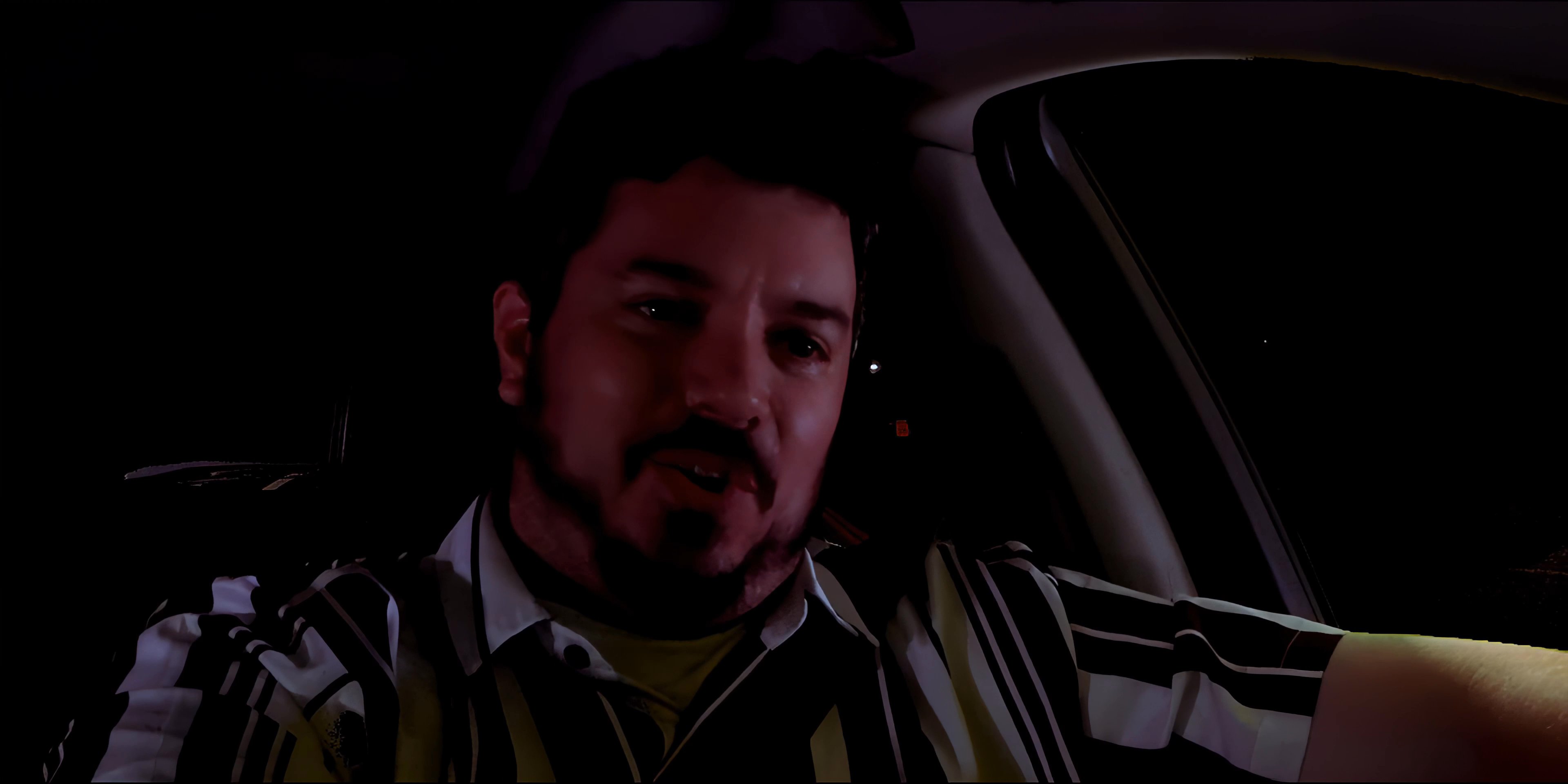 A man engages in a lighthearted conversation while driving his vehicle at night. The sequence captures his expressive facial reactions and his focus on the road through the rearview mirror.