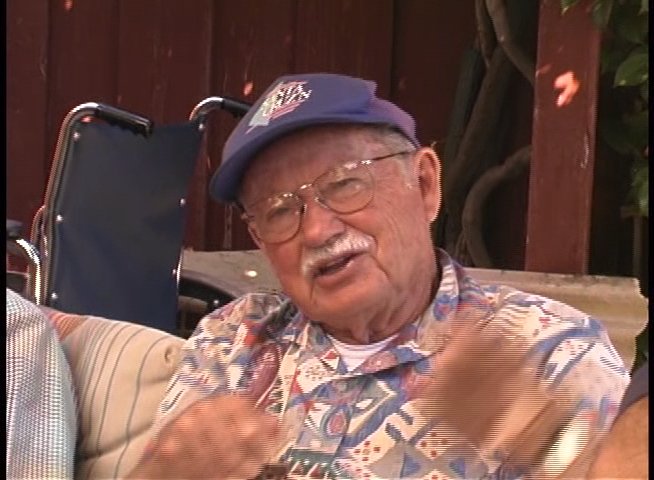 An elderly man shares personal experiences during an outdoor group conversation, occasionally gesturing to emphasize his points. The scene includes an archival photo insert illustrating his historical narrative.