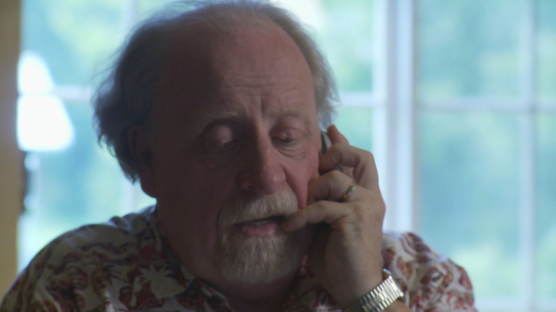 An elderly man with a grey beard engages in a serious, focused telephone conversation while sitting in front of a window.