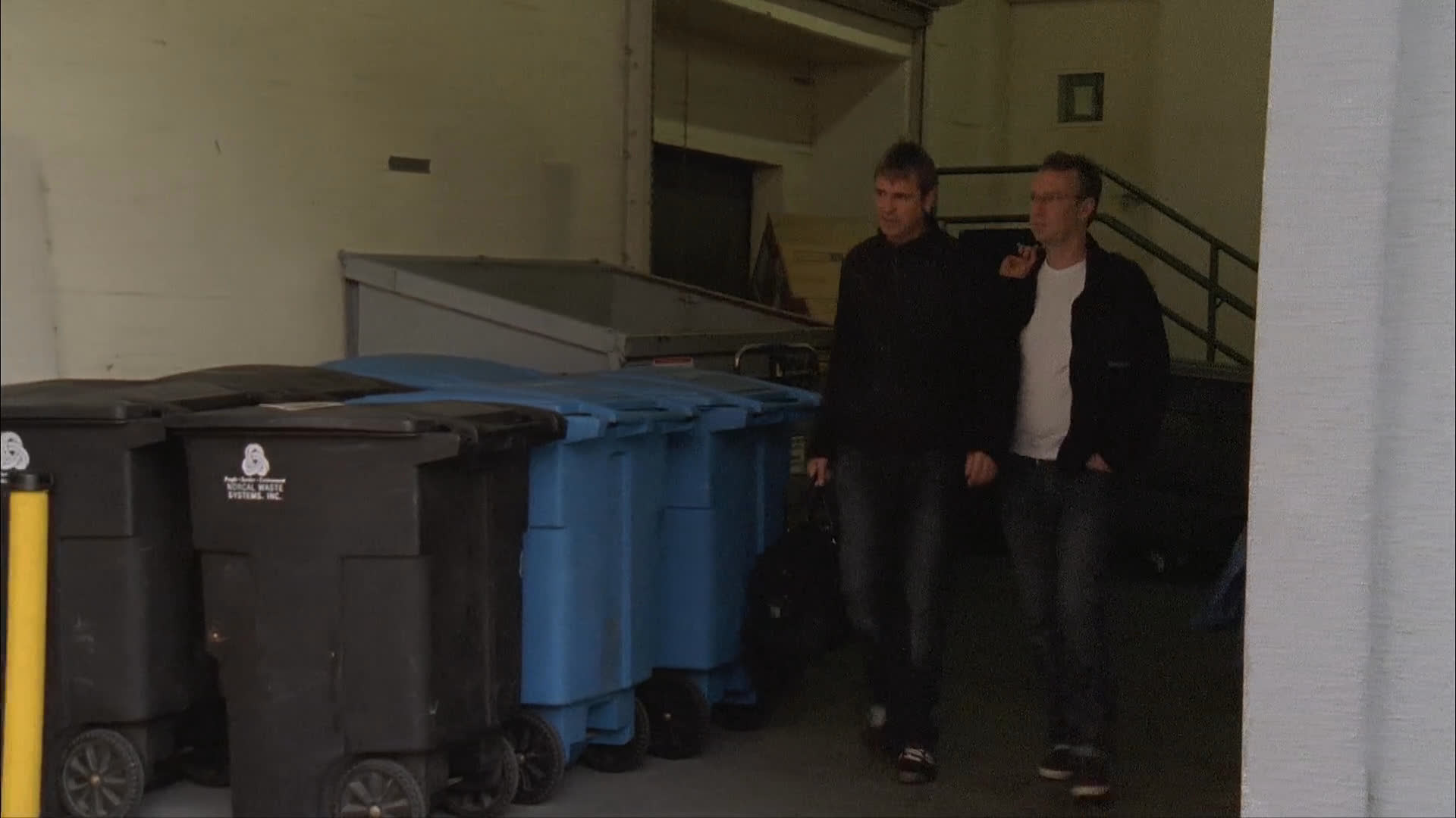 A production team coordinates backstage before transitioning to an exterior urban environment where two men walk along a sidewalk. The sequence concludes with a golden retriever swimming in a body of water.