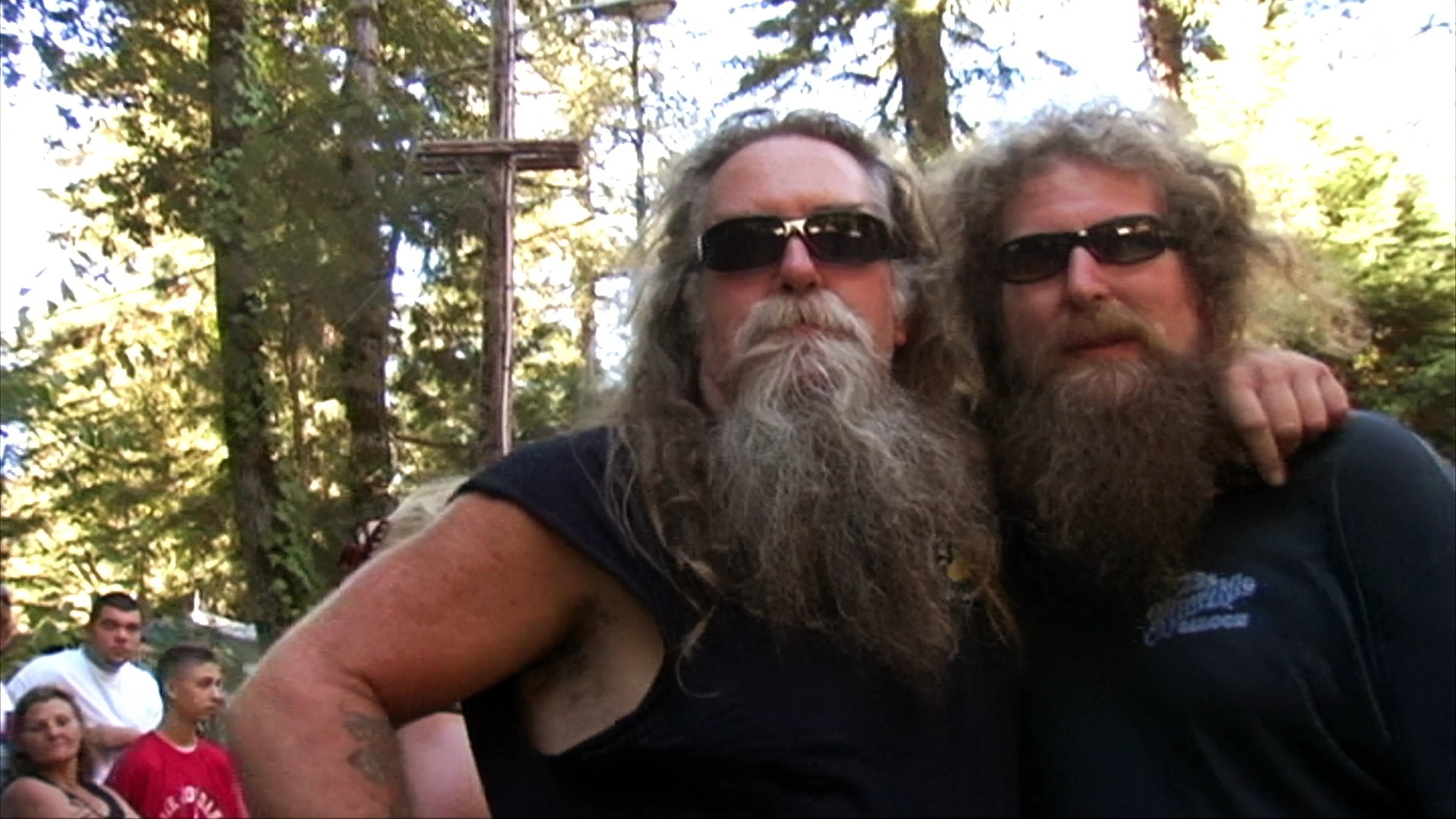 Two bearded men in biker attire interact in an outdoor, woodland setting during a social event. They maintain a friendly demeanor, engaging in casual conversation and gesturing towards one another.
