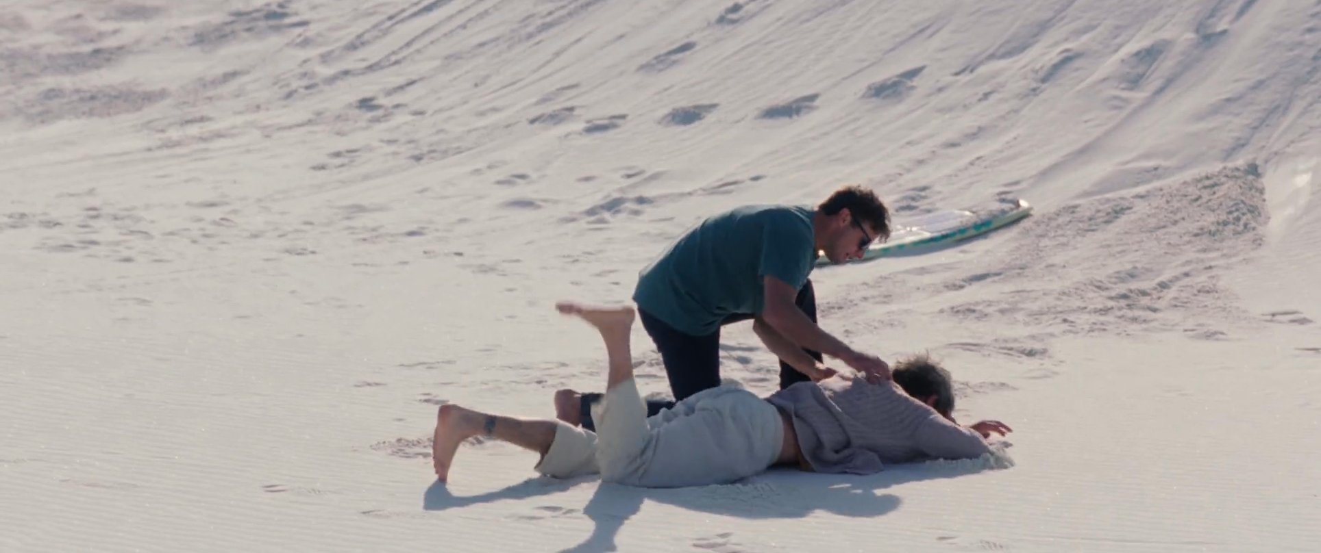 A man loses control while sandboarding down a large dune and suffers a fall, prompting his companion to rush over and assist him.