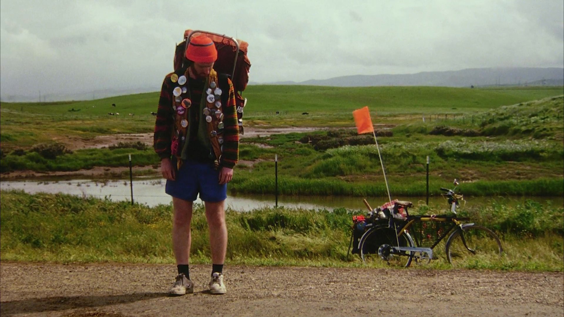 A hiker wearing a heavily accessorized backpack pauses on a roadside during a cross-country journey. The traveler adjusts gear before sitting in a grassy field next to a bicycle.