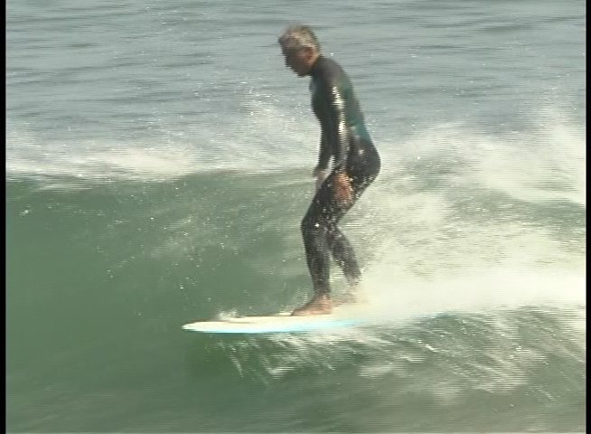 A surfer in a wetsuit skillfully maneuvers a longboard across a breaking ocean wave. The athlete maintains balance and composure while traversing the water surface.