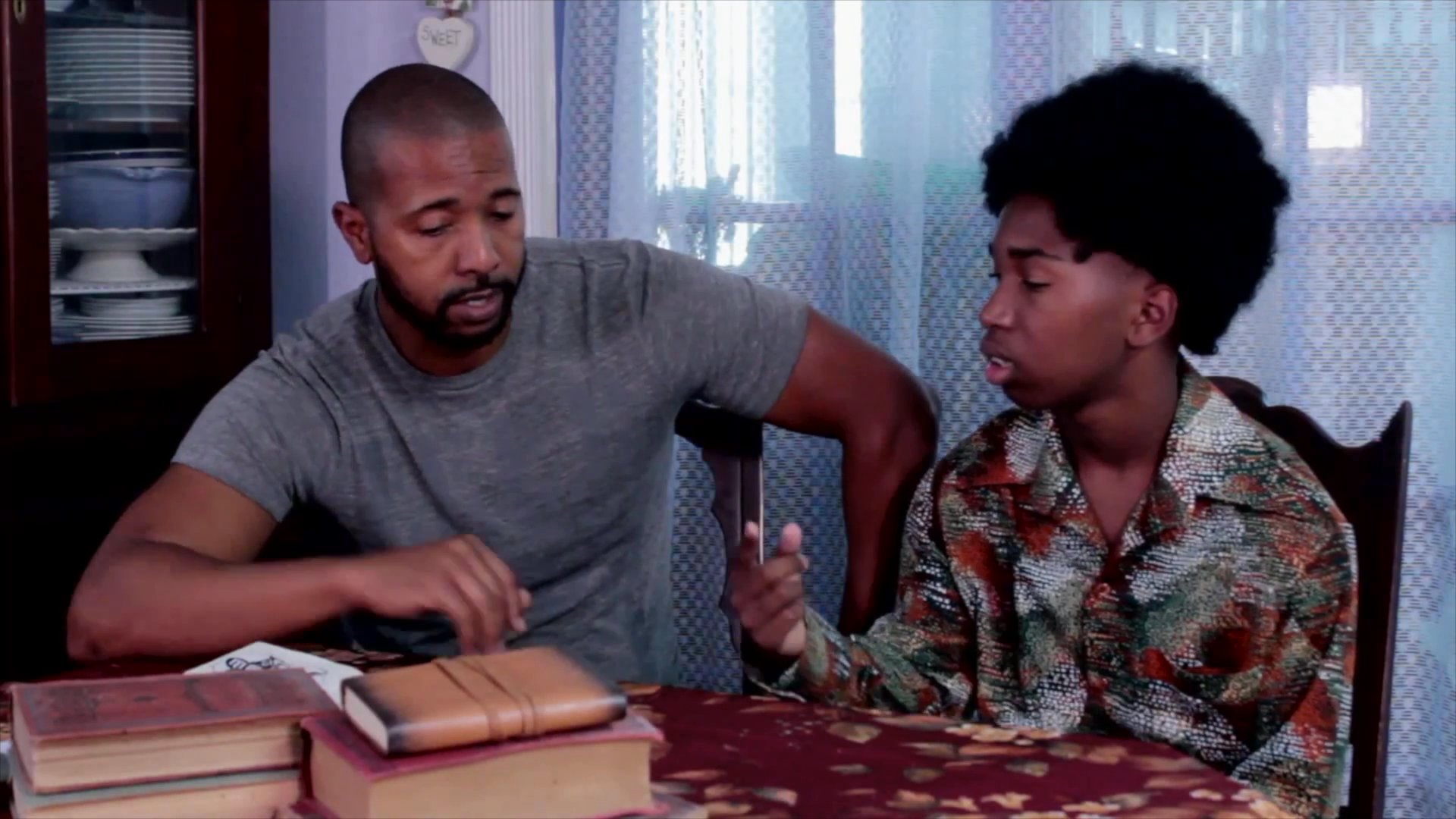 An adult man guides a younger person through historical narratives while reading from an illustrated sketchbook at a kitchen table. They engage in an intimate dialogue about cultural traditions and past events depicted in the handwritten notes and drawings.