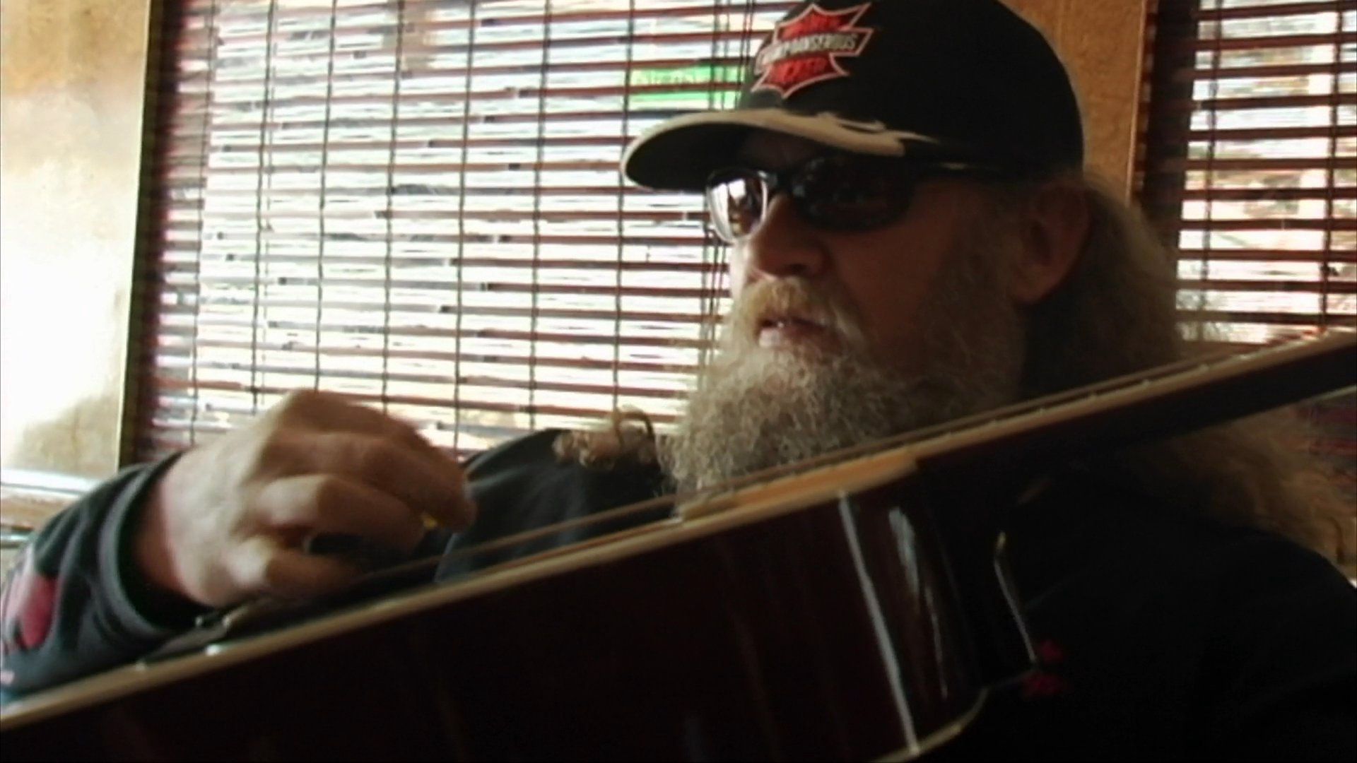 A bearded musician wearing sunglasses and a cap plays an acoustic guitar while sitting indoors near window blinds.