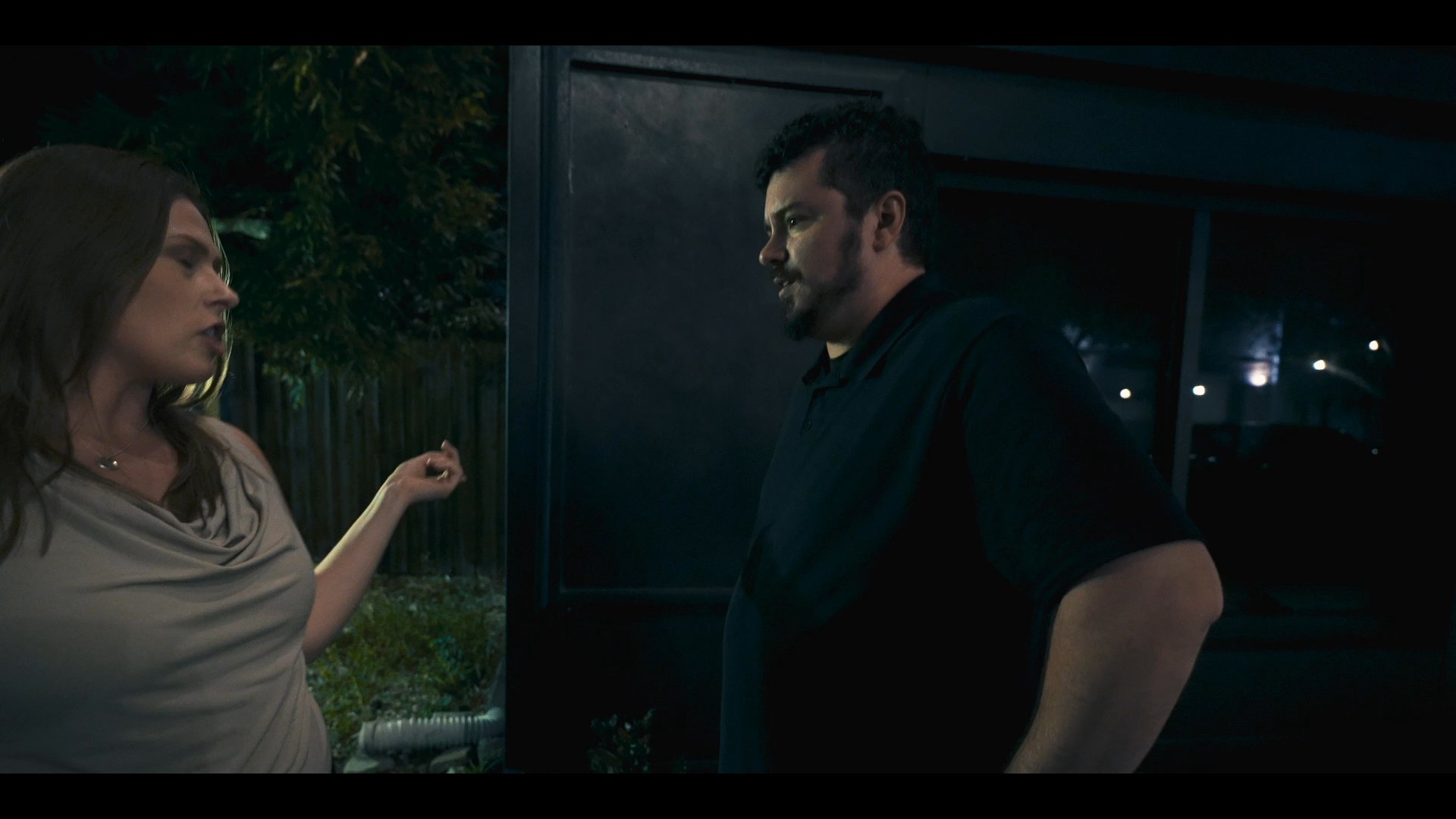 A man and a woman engage in a tense, heated verbal exchange outside a business at night. The interaction appears emotionally charged as they gesture and communicate intensely.