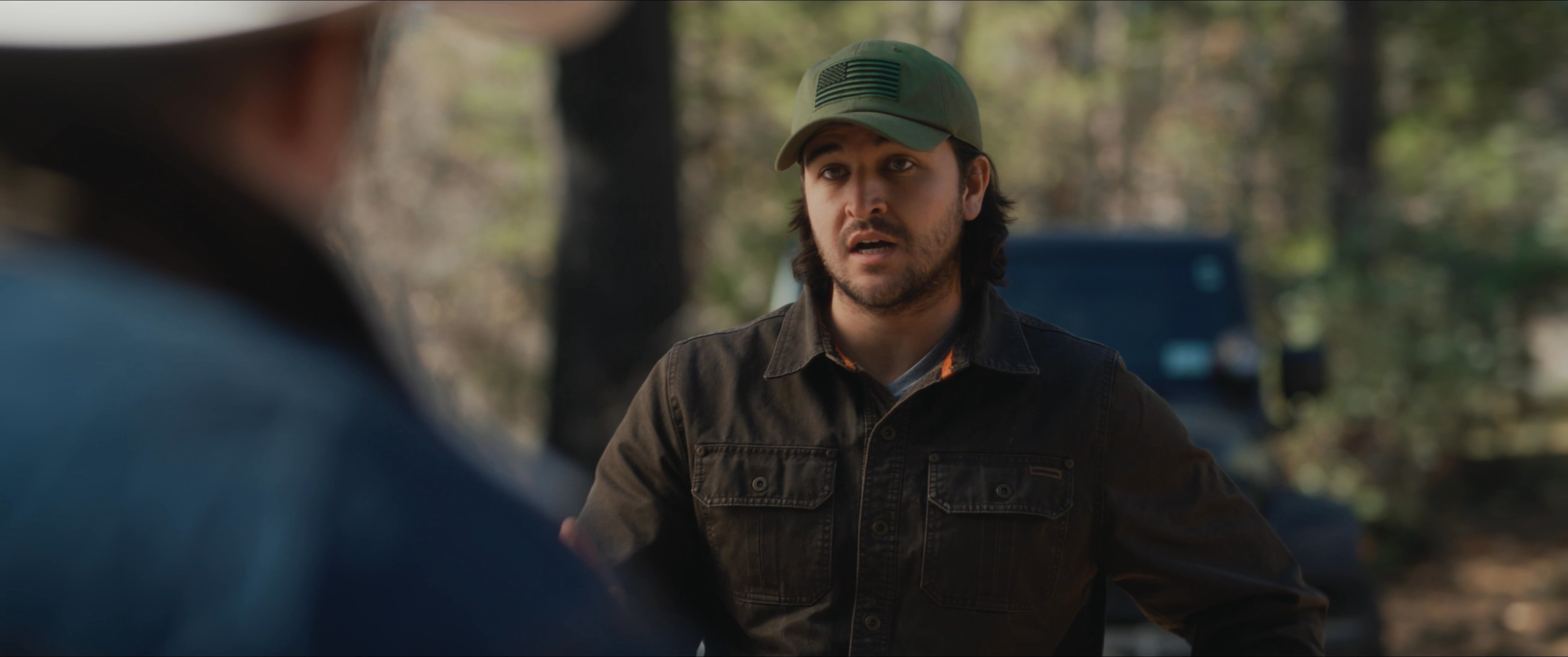 Two men engage in a tense discussion within a wooded clearing, while a third individual observes from a distance. The scene captures an outdoor exchange between characters dressed in rugged attire.