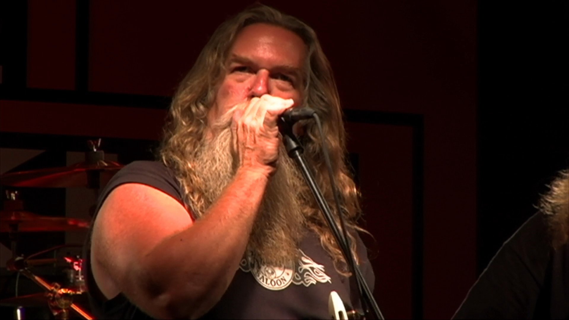 A long-haired, bearded male musician performs vocals on a dimly lit stage at a music venue. The scene transitions to patrons dancing and enjoying the live entertainment in the bar area.