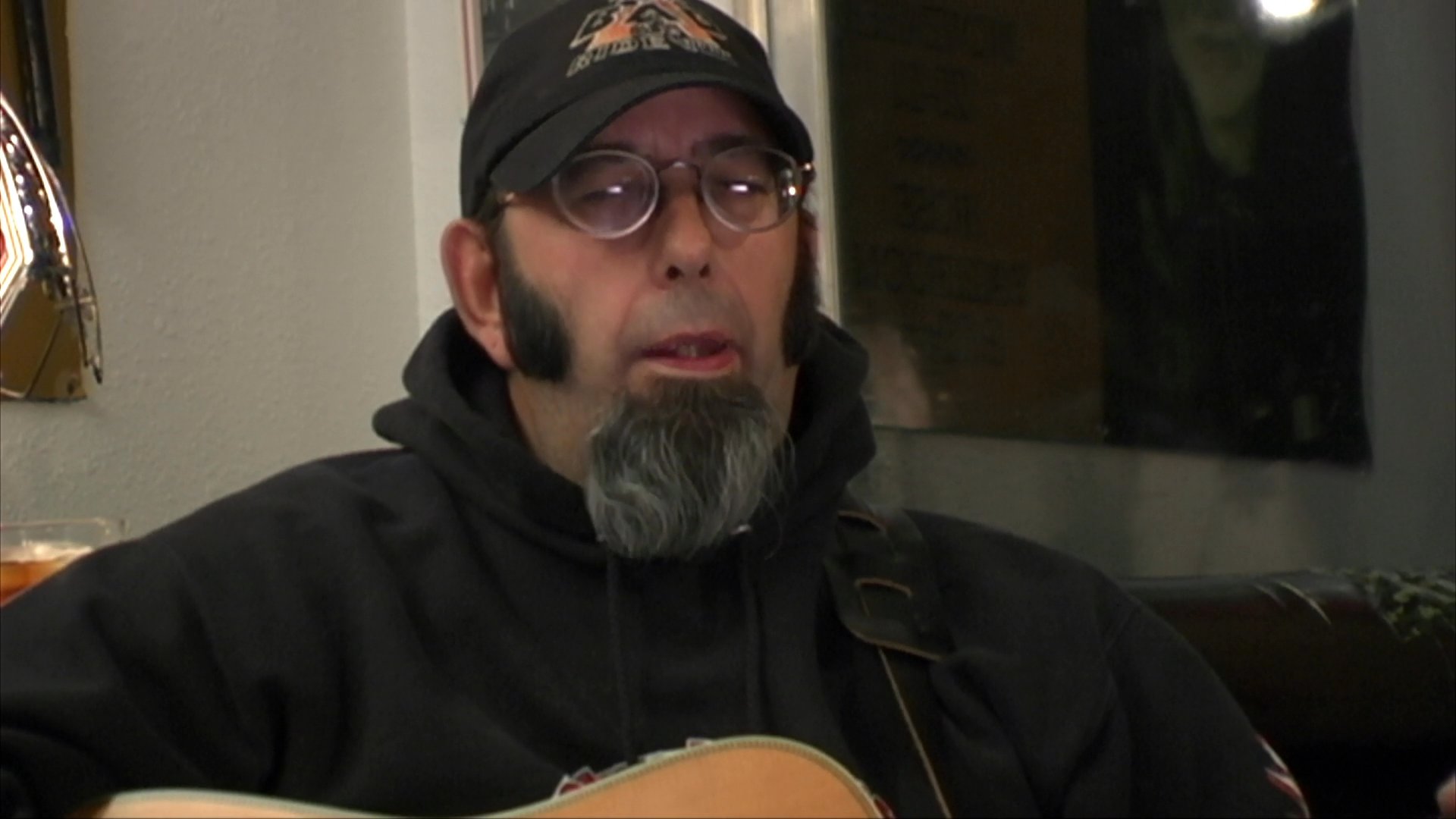 A bearded musician in a hooded sweatshirt performs while playing an acoustic guitar. He sings with focus, maintaining a steady rhythmic engagement throughout the session.