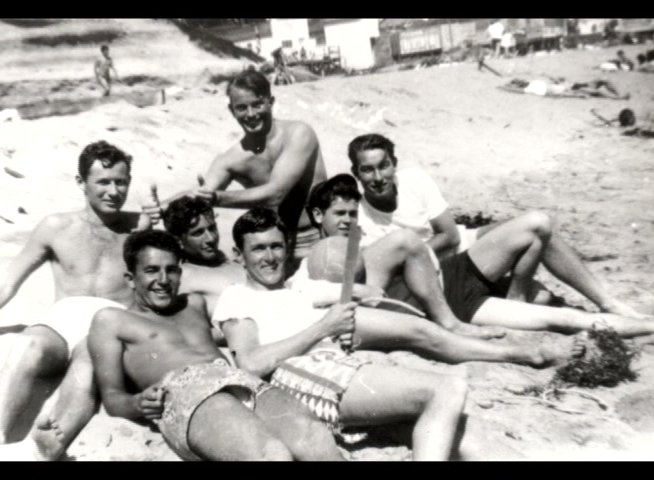An elderly man recounts his youth through personal anecdotes while displaying vintage black-and-white photographs of friends at the beach. The narrative transitions to historical footage of individuals interacting with surfboards on the shore.