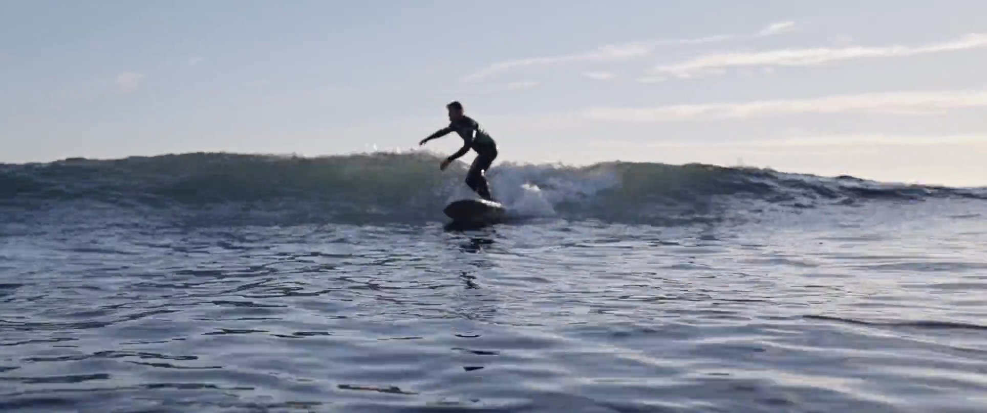A skilled surfer in a wetsuit maneuvers a surfboard across breaking ocean waves against a coastal backdrop.