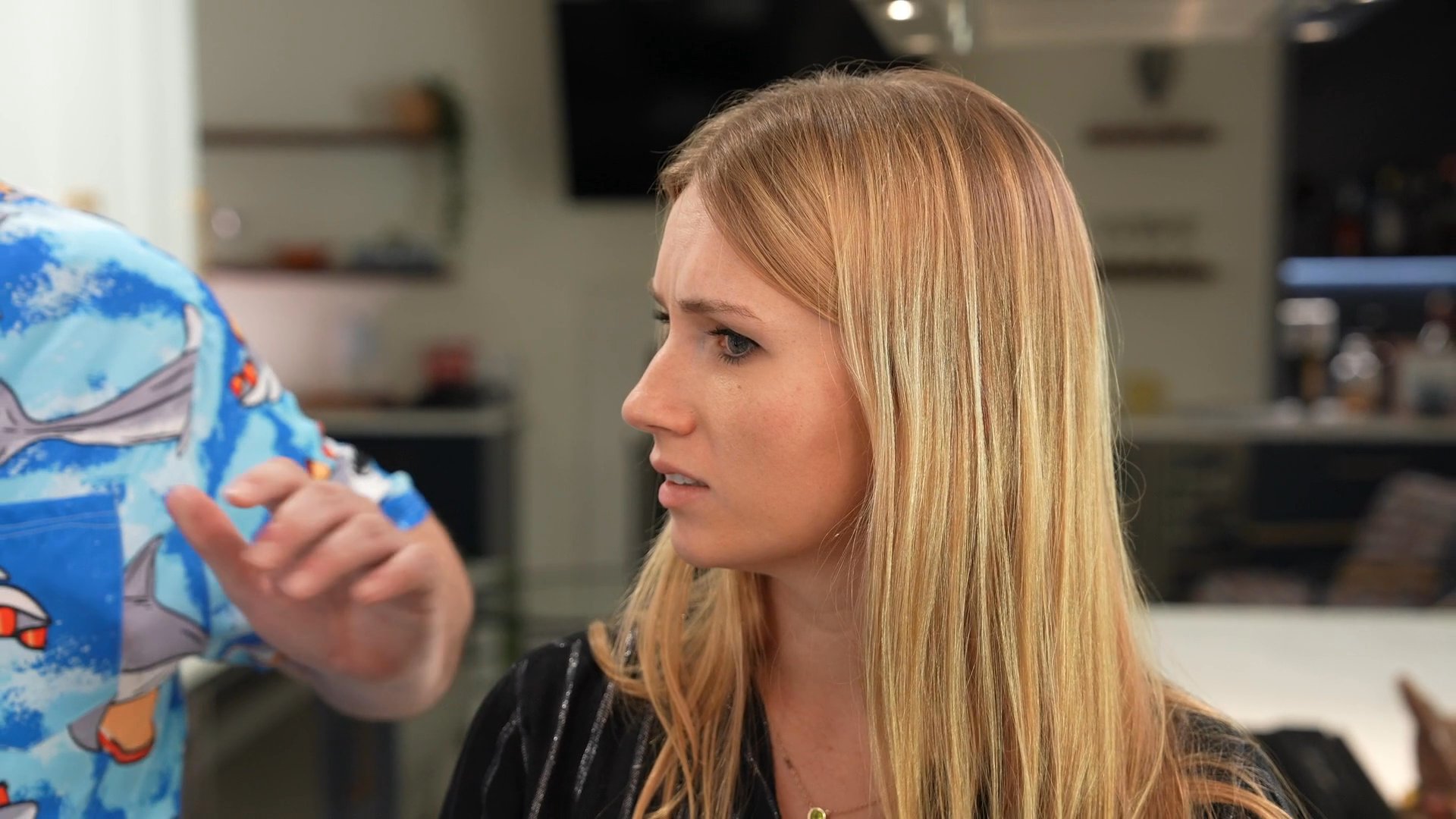 A woman maintains a focused expression while navigating a series of interruptions and conversations during a social gathering at home. The scene transitions to a group discussion involving multiple participants in a kitchen environment.