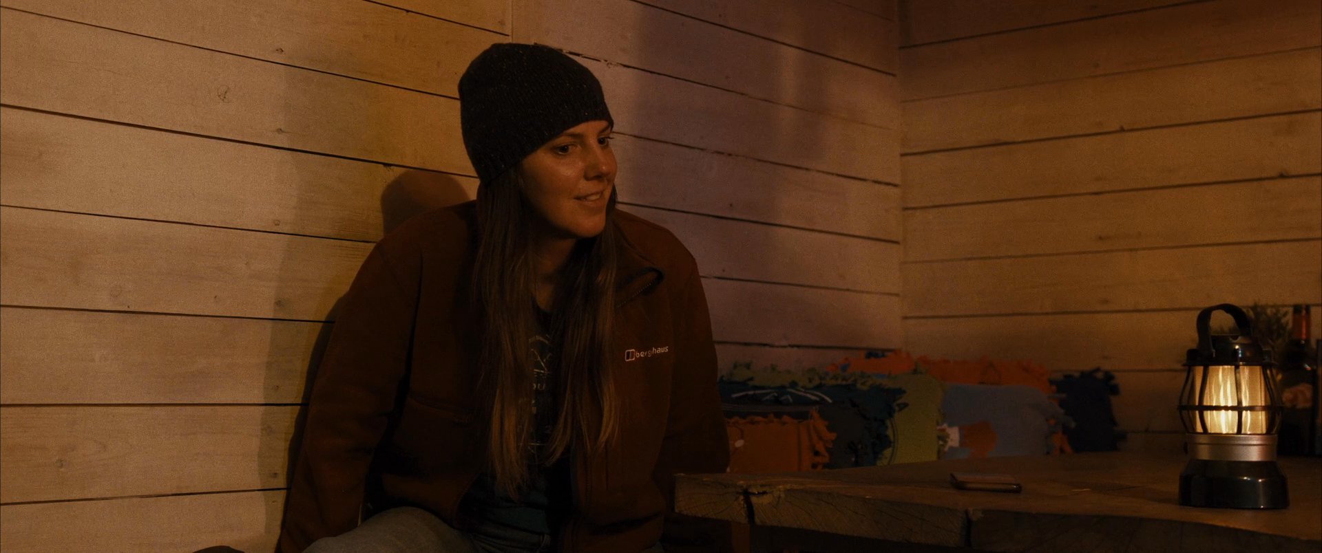Two individuals engage in a quiet dialogue within a dimly lit, wood-paneled cabin illuminated by a lantern and a candle. The atmosphere is intimate and focused as they sit together in the rustic setting.