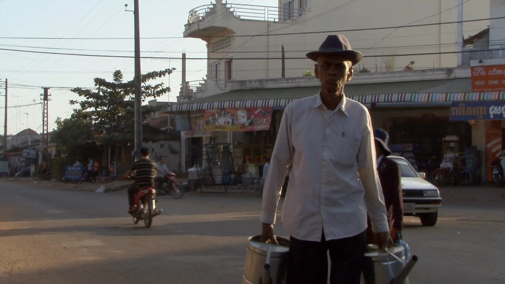 Daily life unfolds on a bustling Cambodian village street, featuring pedestrians, local traffic, and children interacting in the lively outdoor environment.
