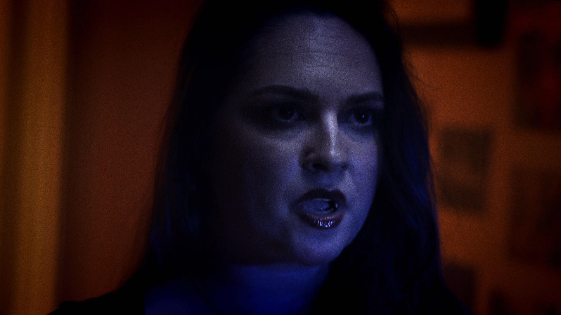 A woman reveals her vampire fangs in a dimly lit, web-covered environment while a man remains bound in the background. The atmosphere conveys a supernatural and eerie narrative tension.