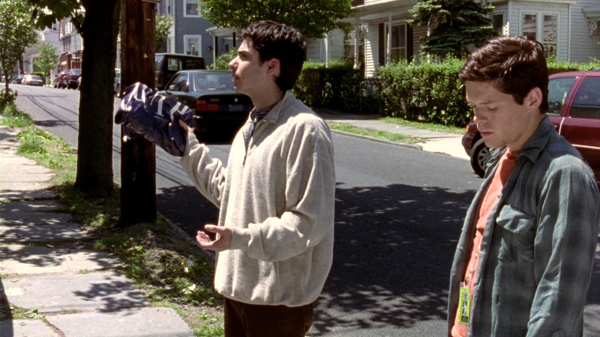 Two individuals engage in a conversation on a suburban residential street, exchanging a small wrapped package before parting ways.