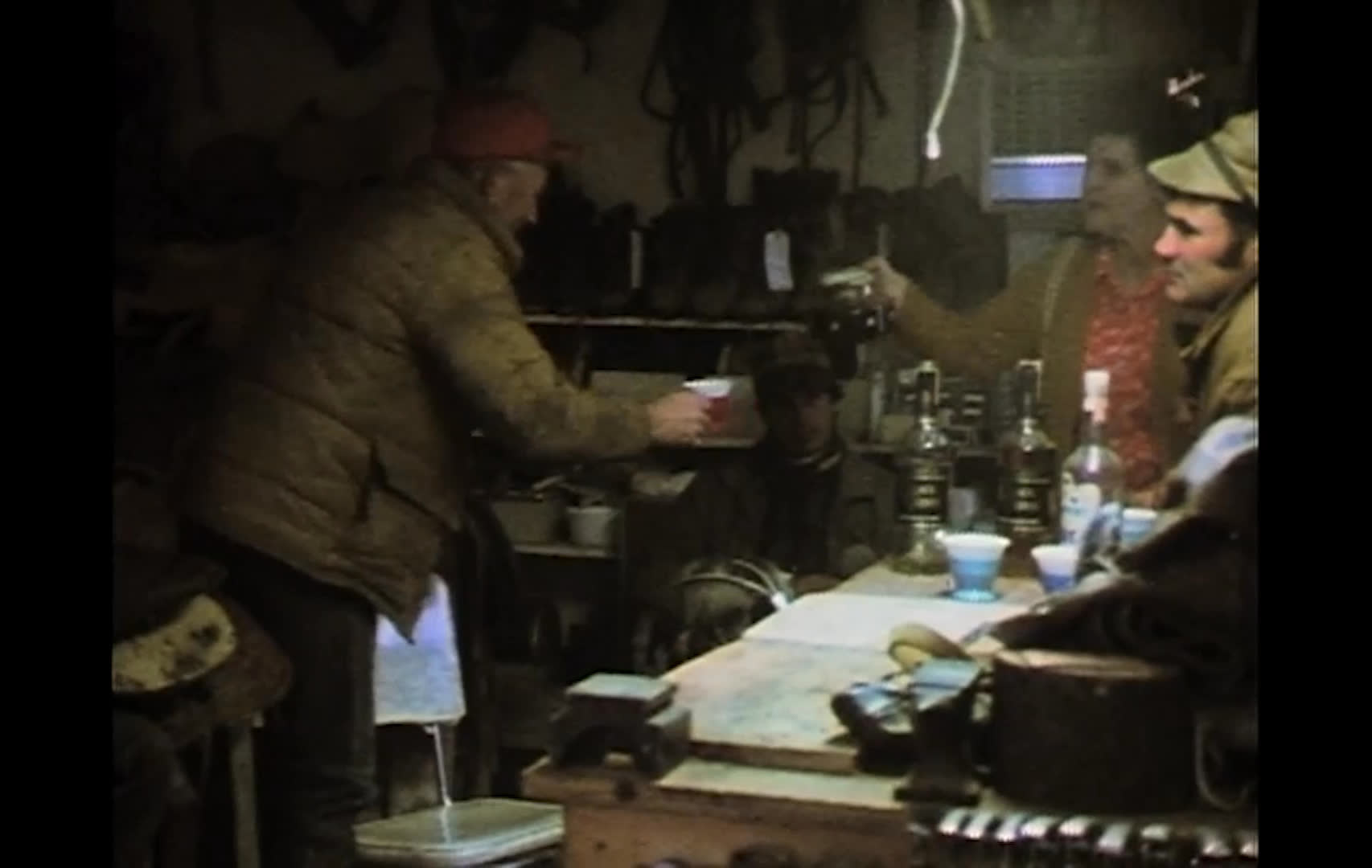 A man enters a weathered saddle shop, where older friends gather among saddles and gear to talk over coffee. One pours from a bottle into a cup as the relaxed conversation continues.