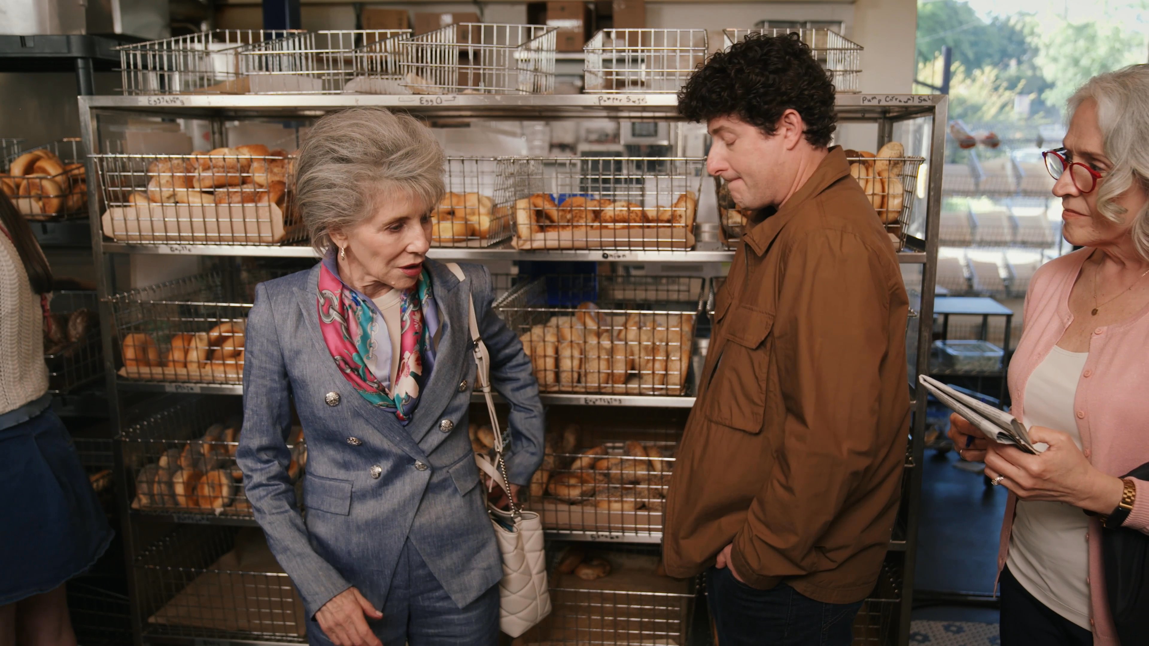 An older woman in a blazer engages in a polite conversation with a male bakery employee near shelves stocked with bagels. They exchange pleasantries while other patrons are visible in the background of the shop.