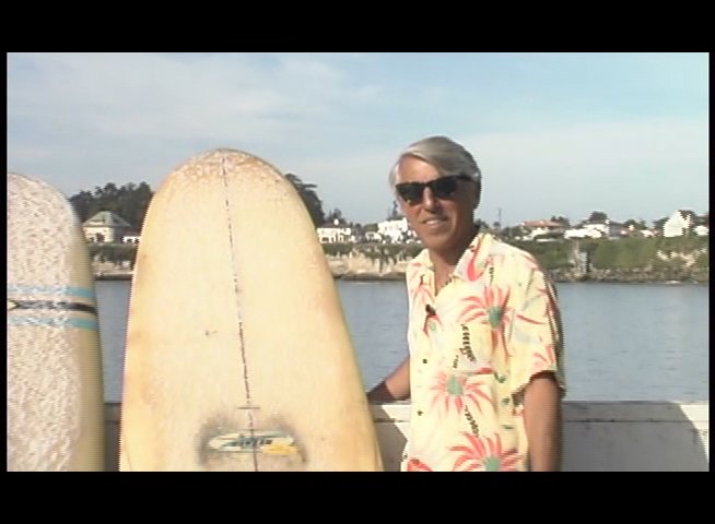 Rene Ouelette presents two surfboards on a coastal railing overlooking a beach with active surfers in the distance.