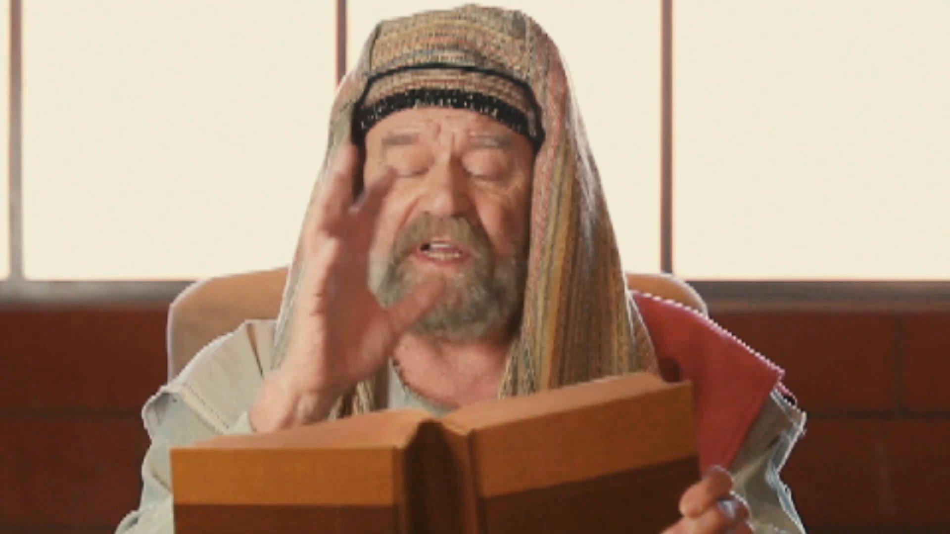 A man dressed in period-appropriate attire recites a story while gesturing expressively from an open book. He maintains an engaging performance style throughout his narration.
