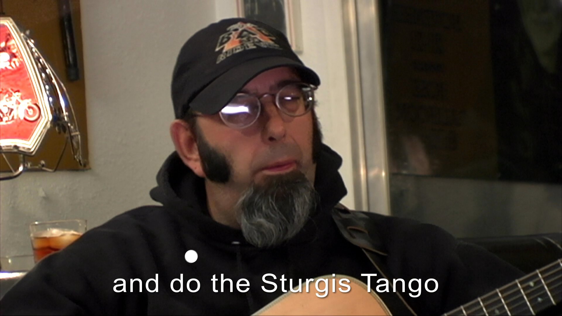 A man wearing a baseball cap performs a song about the Sturgis motorcycle rally while playing an acoustic guitar. The scene contrasts outdoor evening footage of a motorcycle event with an intimate indoor musical performance.