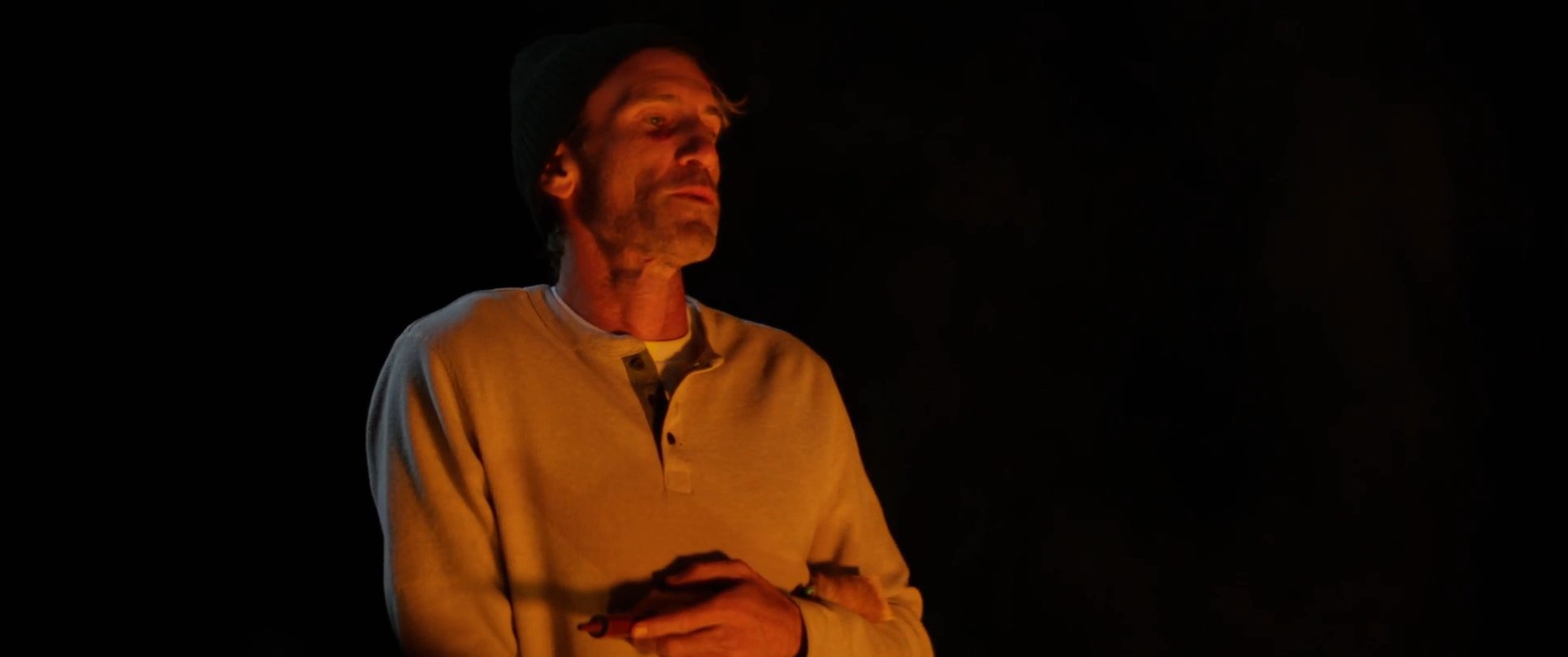 A man shares a story by a campfire while a group listens intently, illuminated by the warm, flickering glow in the dark.