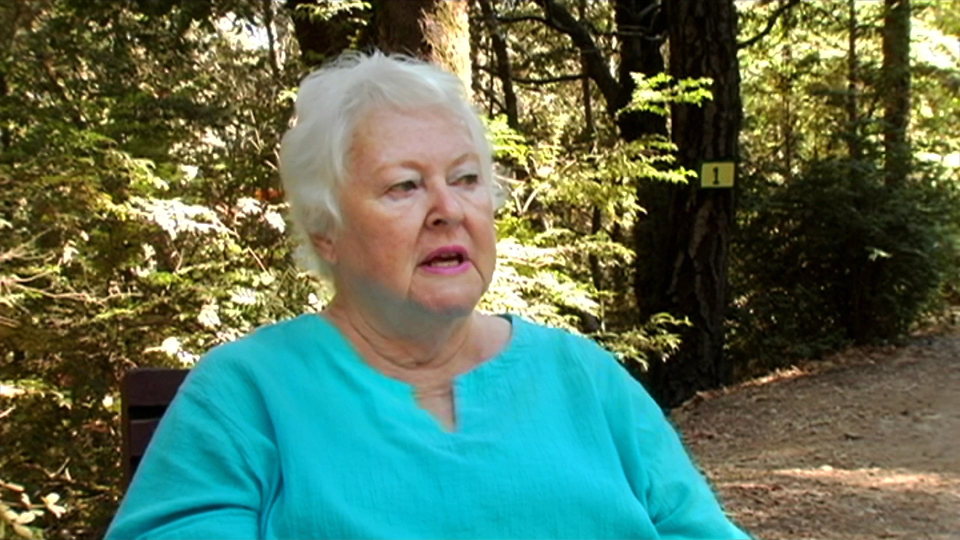 An older woman in a teal blouse engages in a thoughtful conversation while seated in a wooded outdoor environment. She expresses a variety of emotions as she reflects and speaks to an off-camera interviewer.