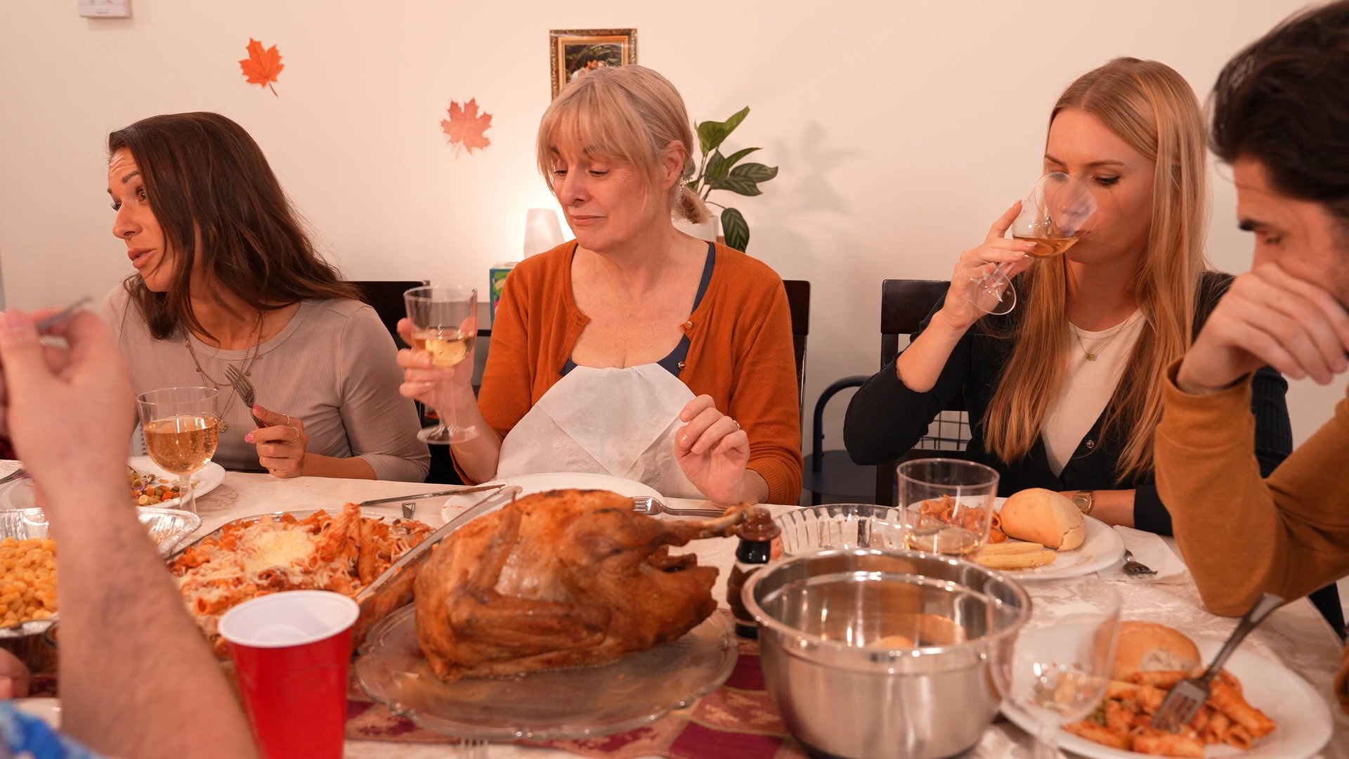 A group of people enjoys a Thanksgiving feast together, sharing conversation and drinks around a table centered with a roasted turkey. The setting reflects a festive home atmosphere as guests interact and dine.